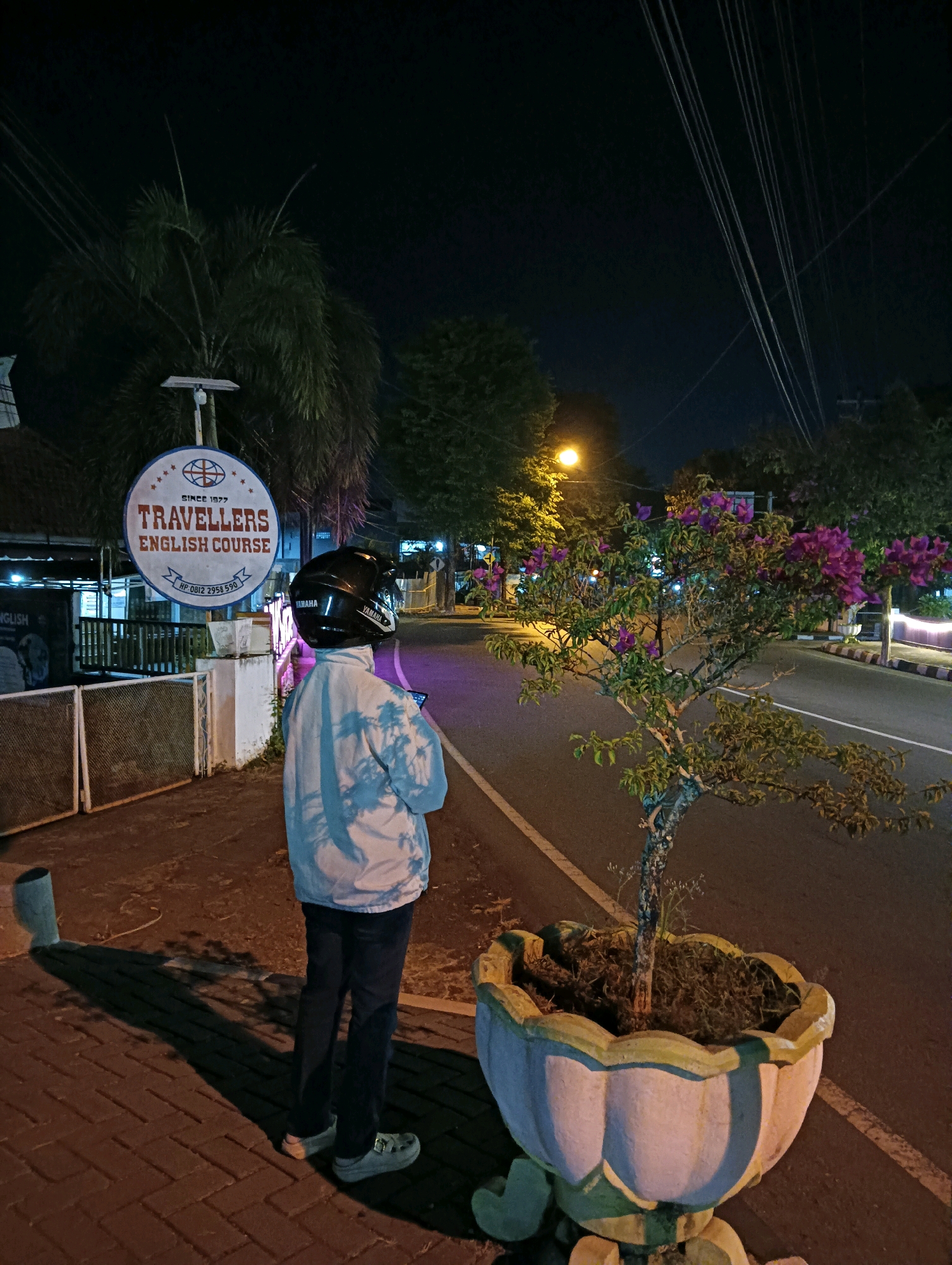 A medium shot shows a person from behind standing on a brick sidewalk at night, looking towards a street lined with trees and a flowering bush. The person is wearing a light-colored, tie-dye jacket, dark pants, and a black helmet with "YAMAHA" branding. They are holding a smartphone. To the left of the person is a sign for "TRAVELLERS ENGLISH COURSE" with contact information below. The environment includes streetlights, power lines, and the shadows of trees cast on the ground. The scene is set in Purworejo, Indonesia, indicated by the context.