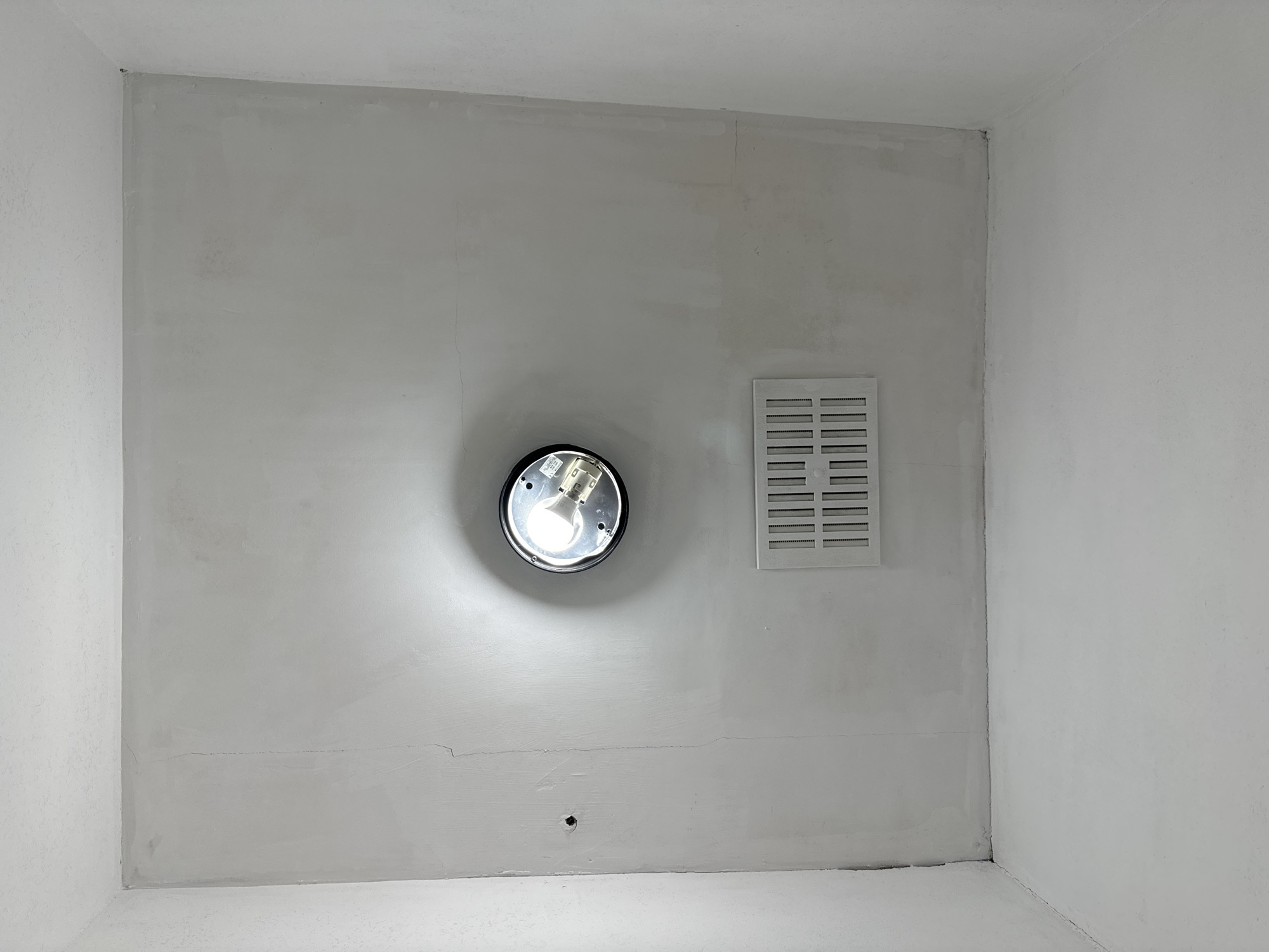 The image shows an indoor scene, possibly a hallway or a room, with white walls and ceiling. In the center of the ceiling, there is a circular light fixture with a visible incandescent bulb. Below the light fixture, a rectangular white vent is installed in the ceiling. The walls appear to have some imperfections, with visible cracks and patches, suggesting recent work or a need for repair. The floor is also white and appears clean. There are no people or any other objects visible in the scene. The overall impression is of a functional but unfinished or slightly neglected interior space. The location in the city of Waterford, Ireland, is not discernible from the visual information provided in the image.