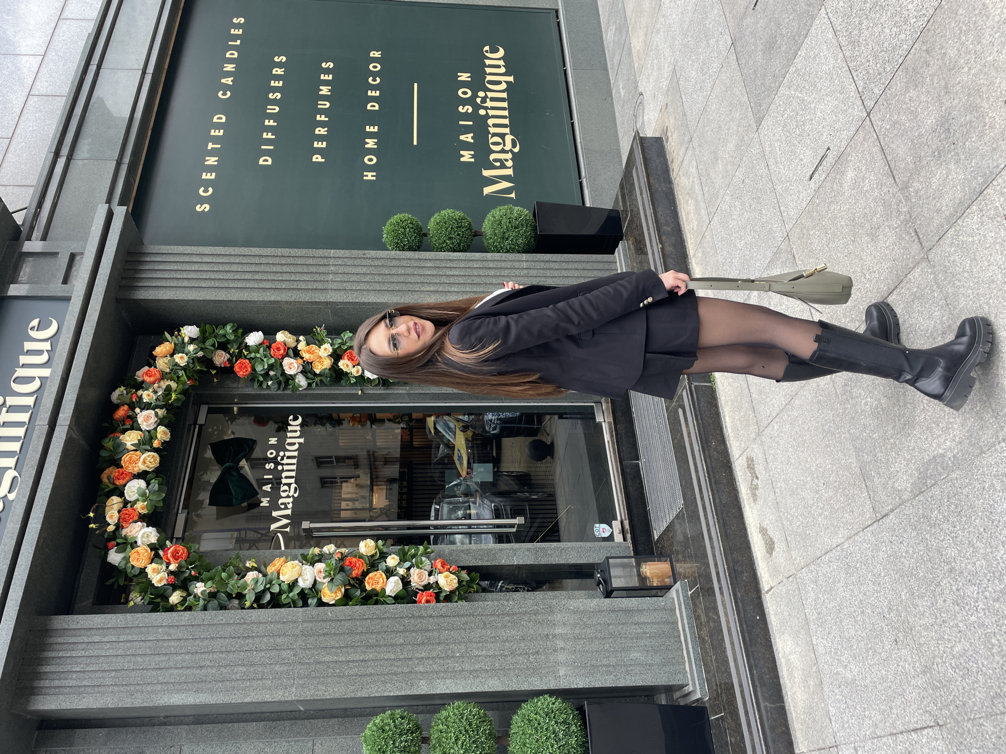 The image shows a woman standing in front of a store named "Maison Magnifique". The store sells scented candles, diffusers, perfumes, and home decor. The storefront is decorated with flowers and topiary plants. The woman is wearing a black blazer, black tights, and black boots. She is holding a green bag. The scene appears to be in an urban environment, and based on the store’s branding, it is likely located in Sofia, Bulgaria. The sidewalk is paved with gray tiles.