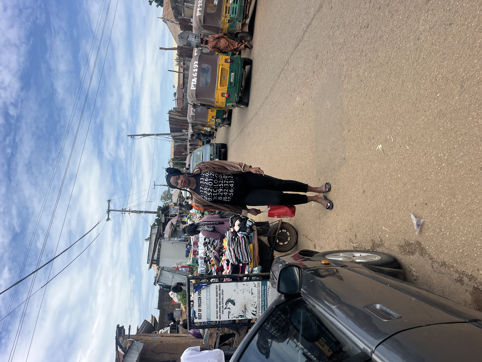 A full shot captures a woman walking down a dusty road in Gura Top, Nigeria. She is wearing a brown bomber jacket with black accents and black leggings, and she carries a red bag in her right hand. Behind her, several auto-rickshaws are parked, some with passengers. The road is lined with buildings, some appearing to be shops or market stalls, with various goods displayed. The sky is cloudy with patches of blue. The scene appears to be a typical street market day.
