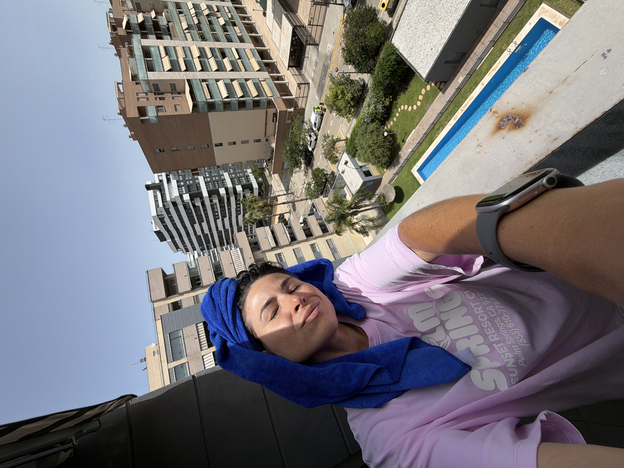 A woman with a blue towel wrapped around her head is standing on a balcony overlooking a city street and several apartment buildings. She is wearing a pink t-shirt with white text that reads "PALM SPRINGS SUNSET RESORT." Sunlight illuminates her face, highlighting freckles. Her eyes are closed, and her head is tilted back. In the background, there is a long, narrow swimming pool with blue water, surrounded by a green lawn and a paved pathway. A few palm trees are visible, along with residential buildings of varying architectural styles and heights. Cars are parked on the street, and there is a fence surrounding a grassy area. The sky is clear and blue.