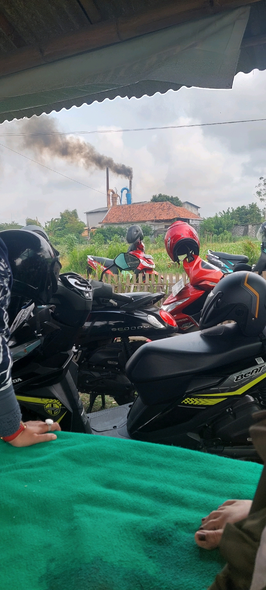In Sumbersuko, Indonesia, a scene unfolds with a focus on parked motorcycles under a rustic, open-air shelter. The shelter's roof, constructed from what appears to be bamboo, casts a shadow over the foreground. Several motorcycles, predominantly scooters, are parked closely together. Helmets rest on some of the bikes, including a black one with orange accents and a red one. In the background, a factory emits dark smoke from a tall chimney, rising into an overcast sky. Green vegetation surrounds the factory, and a wooden fence is visible. A woman's hand, adorned with rings and bracelets, rests on a vibrant green blanket in the lower left of the frame. Her toenails, painted dark, are visible on the blanket in the lower right. The image captures a moment of stillness amidst an industrial backdrop.