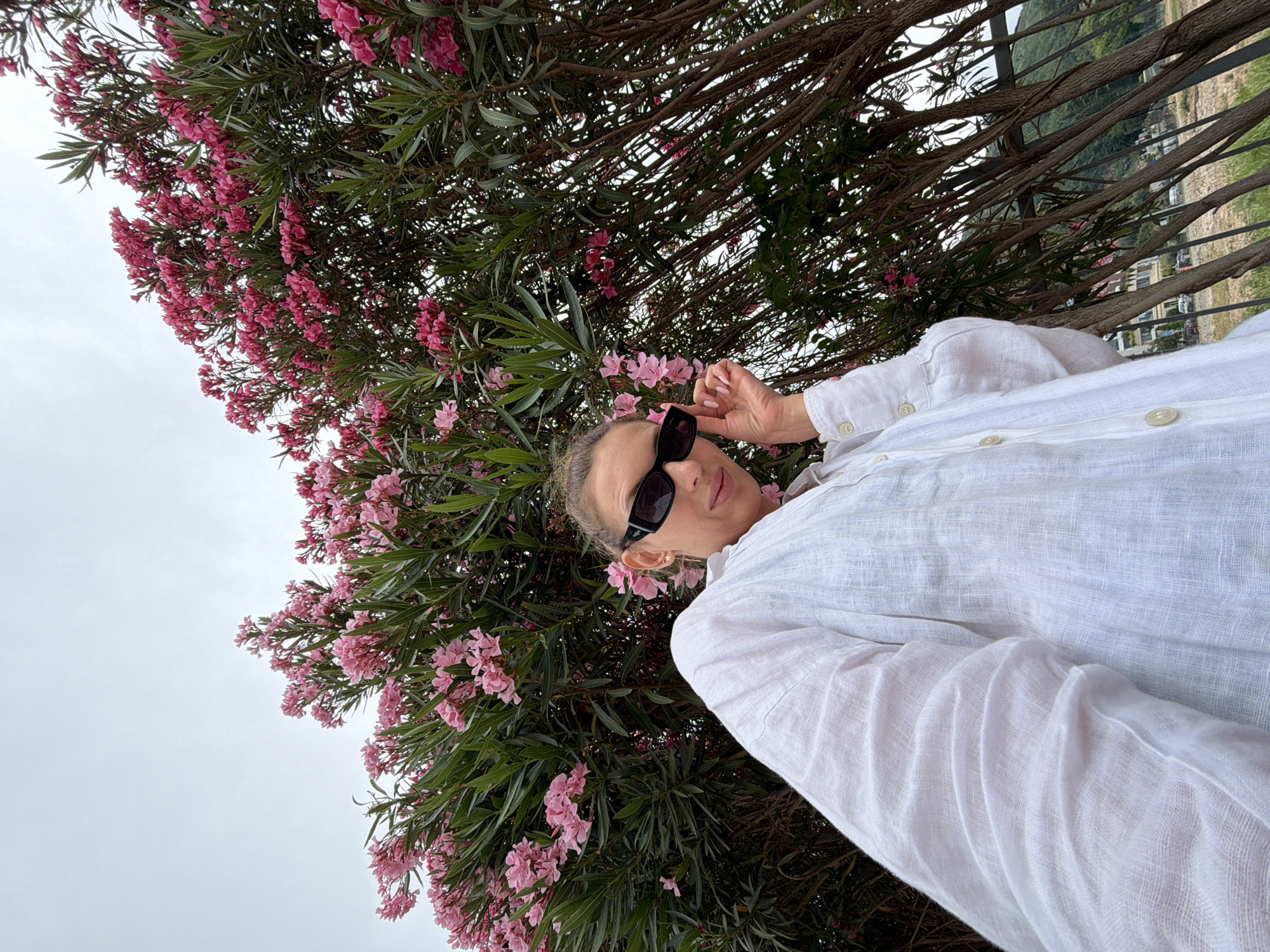 An outdoor, low-angle shot captures a woman posing amidst vibrant pink flowering bushes, likely Oleander, under an overcast sky. The woman, positioned on the right side of the frame, looks directly at the camera while lightly touching her dark sunglasses with her right hand. She is wearing a loose-fitting white shirt, possibly linen. Her hair appears pulled back, and she has light pink nail polish. The dense green foliage and numerous pink blossoms of the bushes dominate the left and central parts of the image, with thick branches forming a natural canopy around her.
In the background, visible through the branches and behind a dark metal railing, is a scenic landscape featuring distant hills, scattered buildings, and what appears to be a body of water. The cloudy, light grey sky fills the upper left portion of the frame. The overall atmosphere suggests a relaxed moment on an overcast day in a coastal or elevated location, consistent with Budva, Montenegro. The woman's expression is neutral, conveying a calm and composed demeanor. No visible text is present.