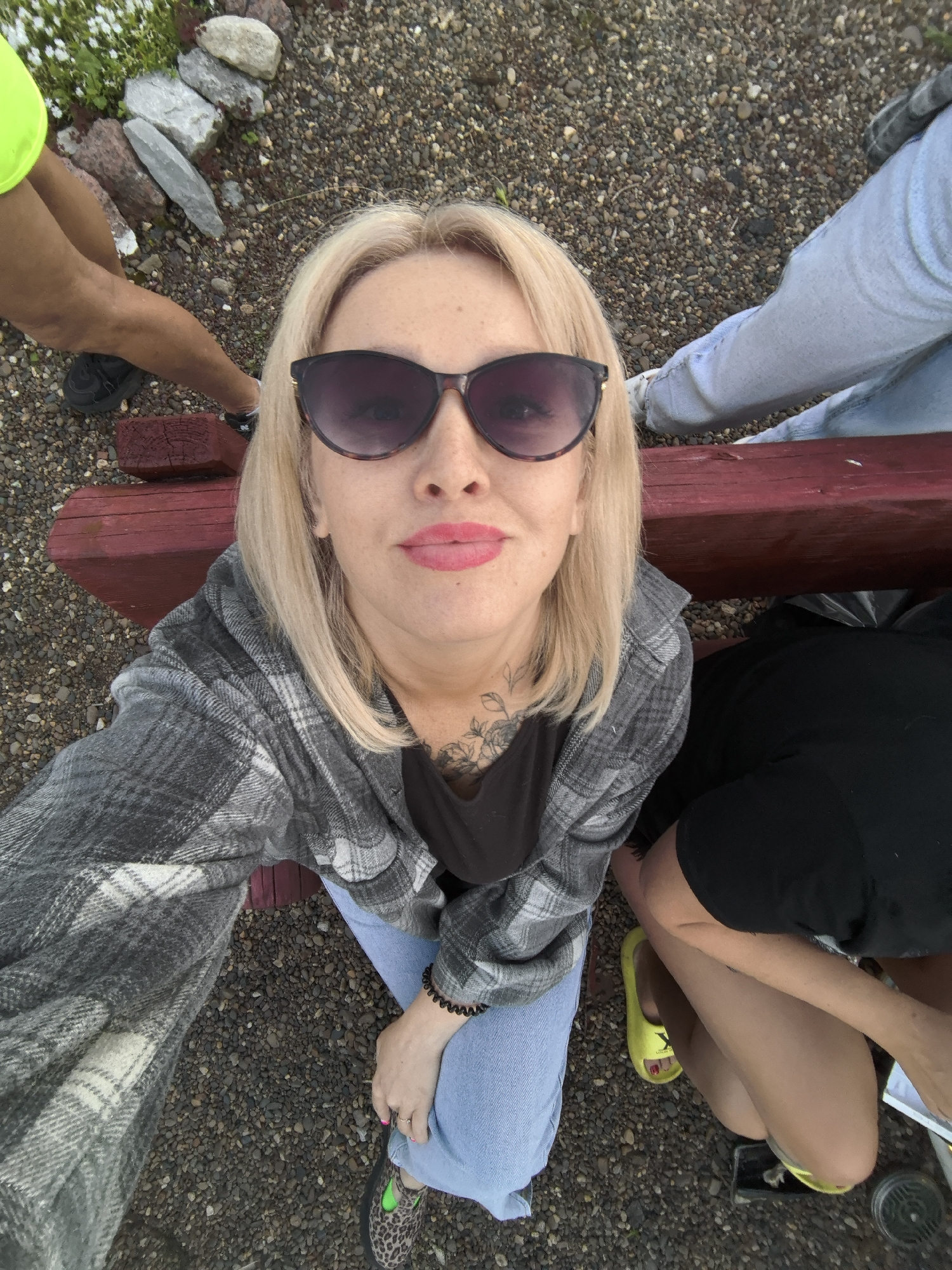 The image is a high-angle selfie taken outdoors, featuring a blonde woman with pink lipstick and dark sunglasses as the primary subject. She wears a grey plaid shirt over a black top and light blue jeans, with a floral tattoo visible on her neck/chest. She is positioned on or next to a red wooden structure, possibly a low bench or steps.
Around her, the legs and arms of at least three other individuals are partially visible, suggesting a casual gathering in Noviy, Russia. To the left, a person is seen in bright neon yellow attire and dark sneakers. To the right, another individual wears light blue jeans and white sneakers. Further down on the right, a third person wears a black top and distinctive yellow slides with a Y-shaped logo (likely Yeezy).
The ground is covered with dark pebbles and gravel, with a small patch of green vegetation and white flowers visible in the top left corner, alongside a small pile of rocks. The lighting indicates it is daytime, with soft, indirect illumination suggesting an overcast sky. The overall scene depicts a relaxed, informal outdoor social setting.