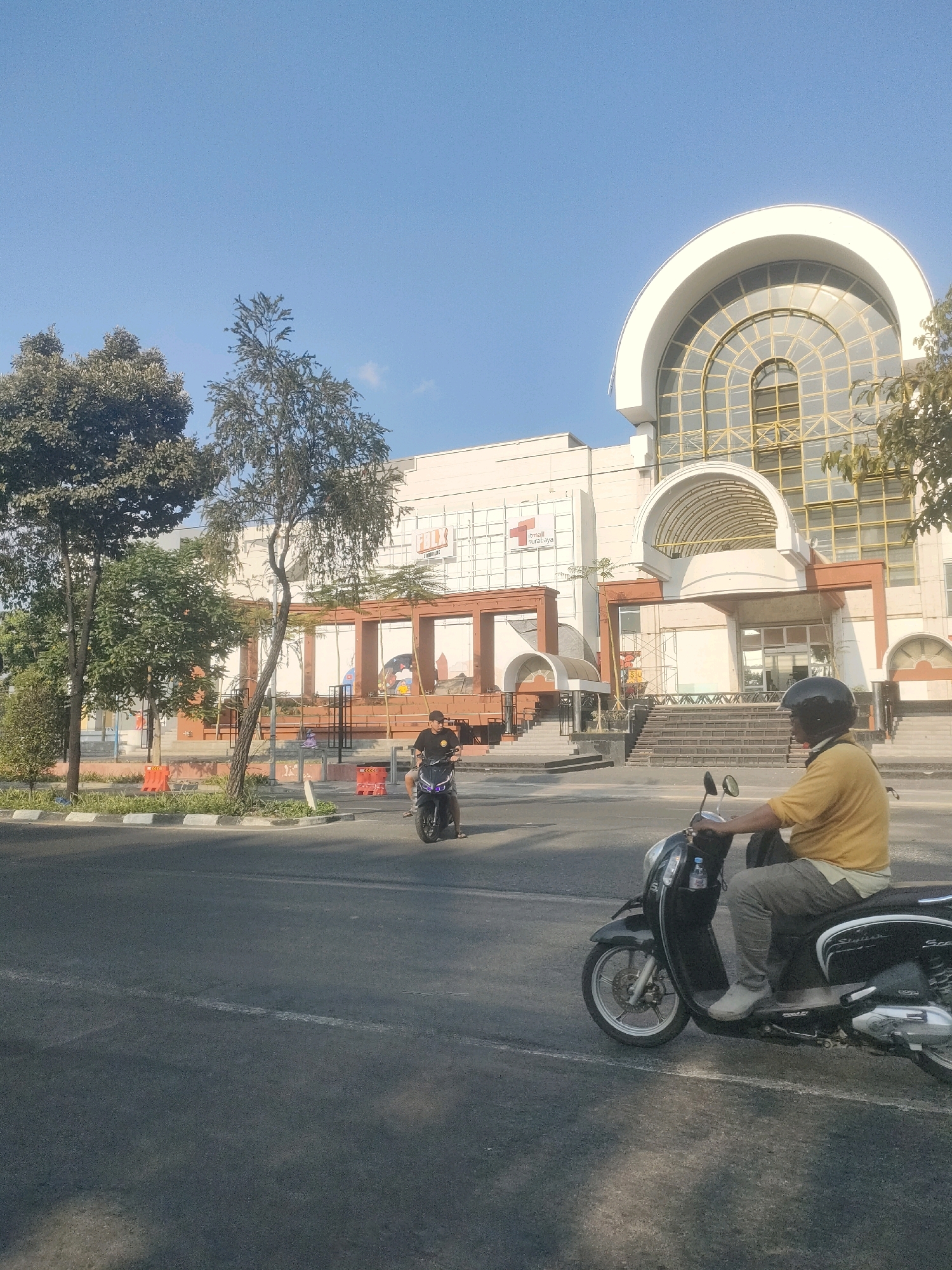 The image depicts a street scene in Surabaya, Indonesia, featuring a modern building with a large arched glass facade. A person wearing a yellow shirt and helmet rides a black scooter in the foreground, facing away from the camera. To the left, another person on a motorcycle is visible near the entrance of the building. The building appears to be a commercial establishment, possibly a mall, with signage indicating "iTmall Surabaya". The street is paved and a sidewalk with greenery lines the building's edge. The sky is clear and blue, suggesting a sunny day.
