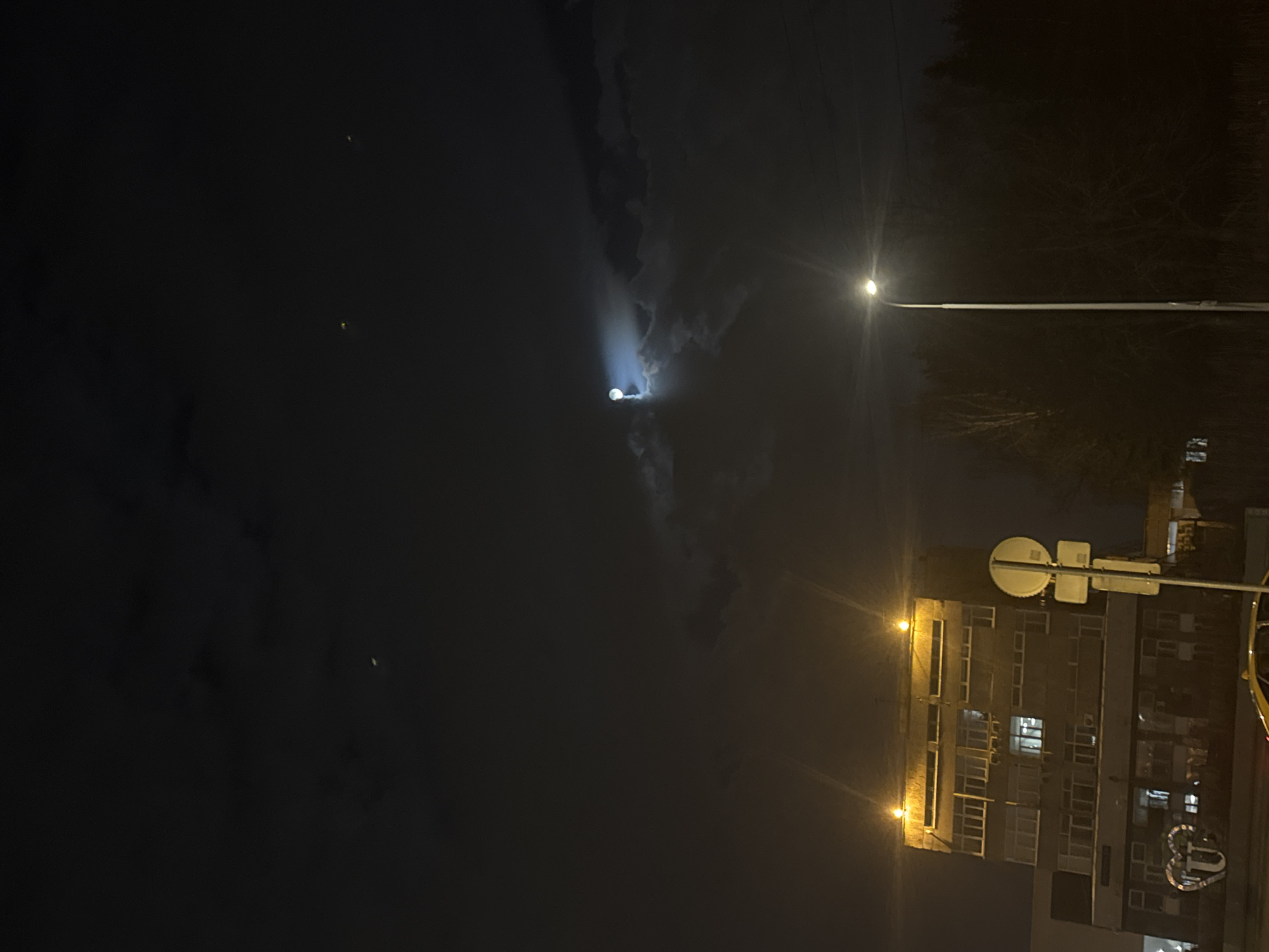 The image depicts a nighttime urban scene in Moscow, Russia. A tall apartment building with multiple illuminated windows is visible on the right side of the image. Two streetlights shine, one near the building and another further away against a dark backdrop of trees or a park. A bright light, possibly from a spotlight or another source of illumination, is directed upwards into the night sky. Dark clouds fill a large portion of the sky, with a few faint pinpricks of light above and to the left. The image appears to have been rotated counter-clockwise by 90 degrees.