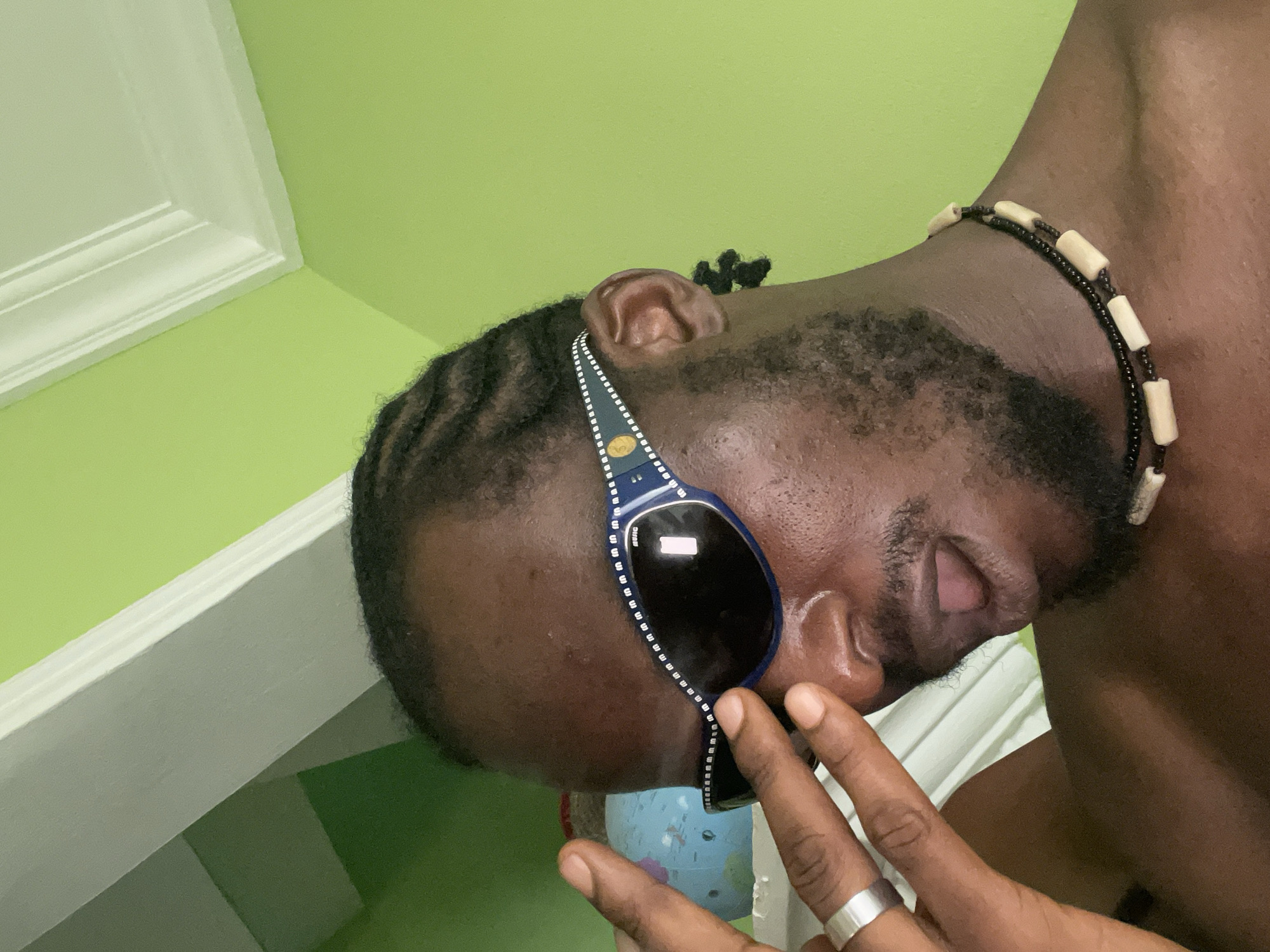 The image is a close-up, high-angle shot of a dark-skinned person wearing sunglasses and a beaded necklace. The person has short, braided hair and a beard. The environment appears to be indoors, with a light green wall and white trim in the background. A small globe is partially visible in the lower-left corner. The person's right hand is raised, with a silver ring visible on one of the fingers.