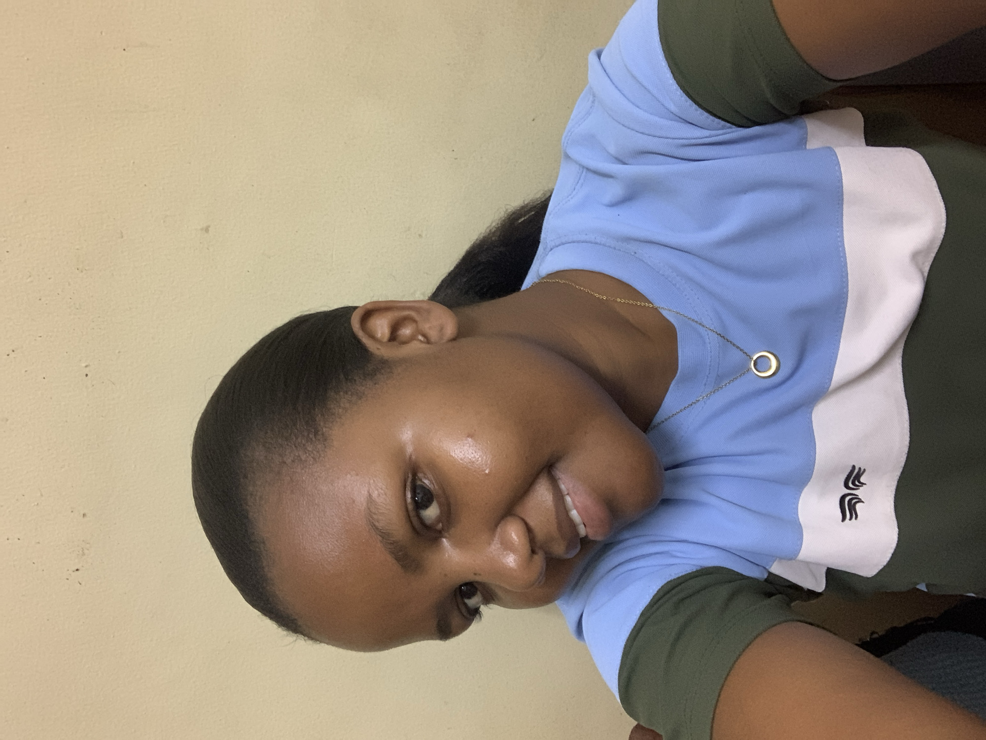 A close-up portrait, oriented 90 degrees clockwise, features a young Black woman smiling warmly at the camera. The setting, provided as Dar es-Salaam, Tanzania, shows a plain, light beige or cream-colored wall as the background, exhibiting minor textural variations and faint marks.
The woman has dark skin and her hair is pulled back, with dark strands visible. She is wearing a collared polo shirt with a distinctive color block design: the shoulders and lower section are olive green, while the main body is light blue, separated by a horizontal white stripe across the chest. On this white stripe, a small, stylized black logo, resembling three wavy lines, is visible. She accessorizes with a delicate gold chain necklace featuring a small, circular gold pendant.
Her facial expression is pleasant and inviting, characterized by a broad smile that shows her teeth. Her dark eyes are bright, and there's a subtle sheen on her forehead and cheeks, suggesting soft lighting. The image captures a moment of direct engagement with the viewer, conveying an amiable demeanor. No specific time of day or weather conditions are discernible from this indoor portrait.