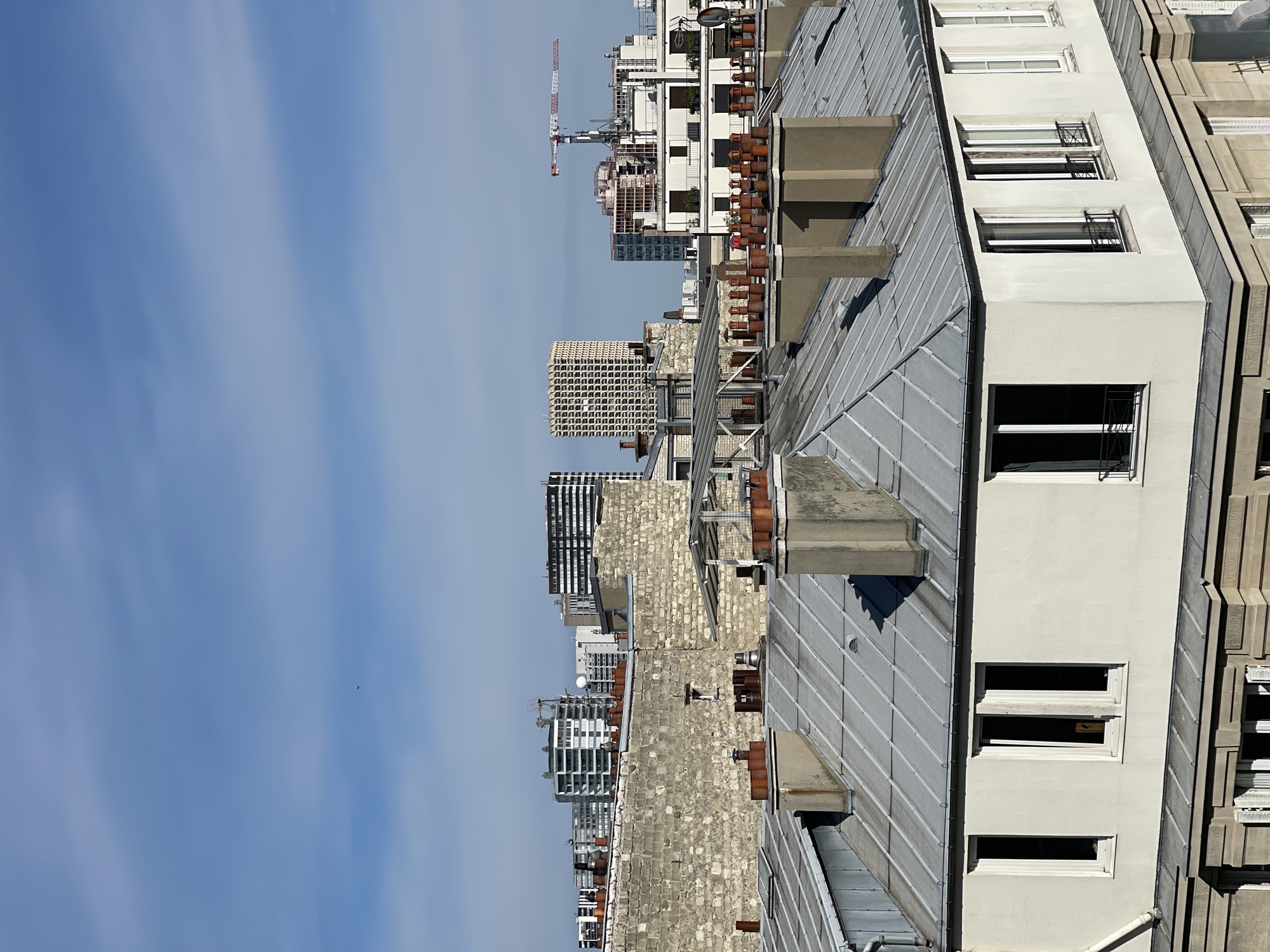 This media file depicts a rooftop view of a cityscape, likely Paris, France, given the architectural style. The scene is dominated by the rooftops of several buildings, varying in height and architectural design. The closest building features a light-colored facade with several windows, some with protective metal bars. Its roof is covered in metal sheeting. Adjacent to it, another building's roof is made of similar metal, with visible chimney pots and concrete structures. Further in the background, a variety of other buildings are visible, including some with brick facades and more modern, glass-fronted structures. A construction crane stands tall in the distance, indicating ongoing development. The sky is a clear, pale blue with subtle cloud wisps, suggesting a daytime setting with fair weather. No people or specific events are visible; the focus is on the urban landscape. The overall impression is one of an urban environment under a bright, possibly sunny, day.