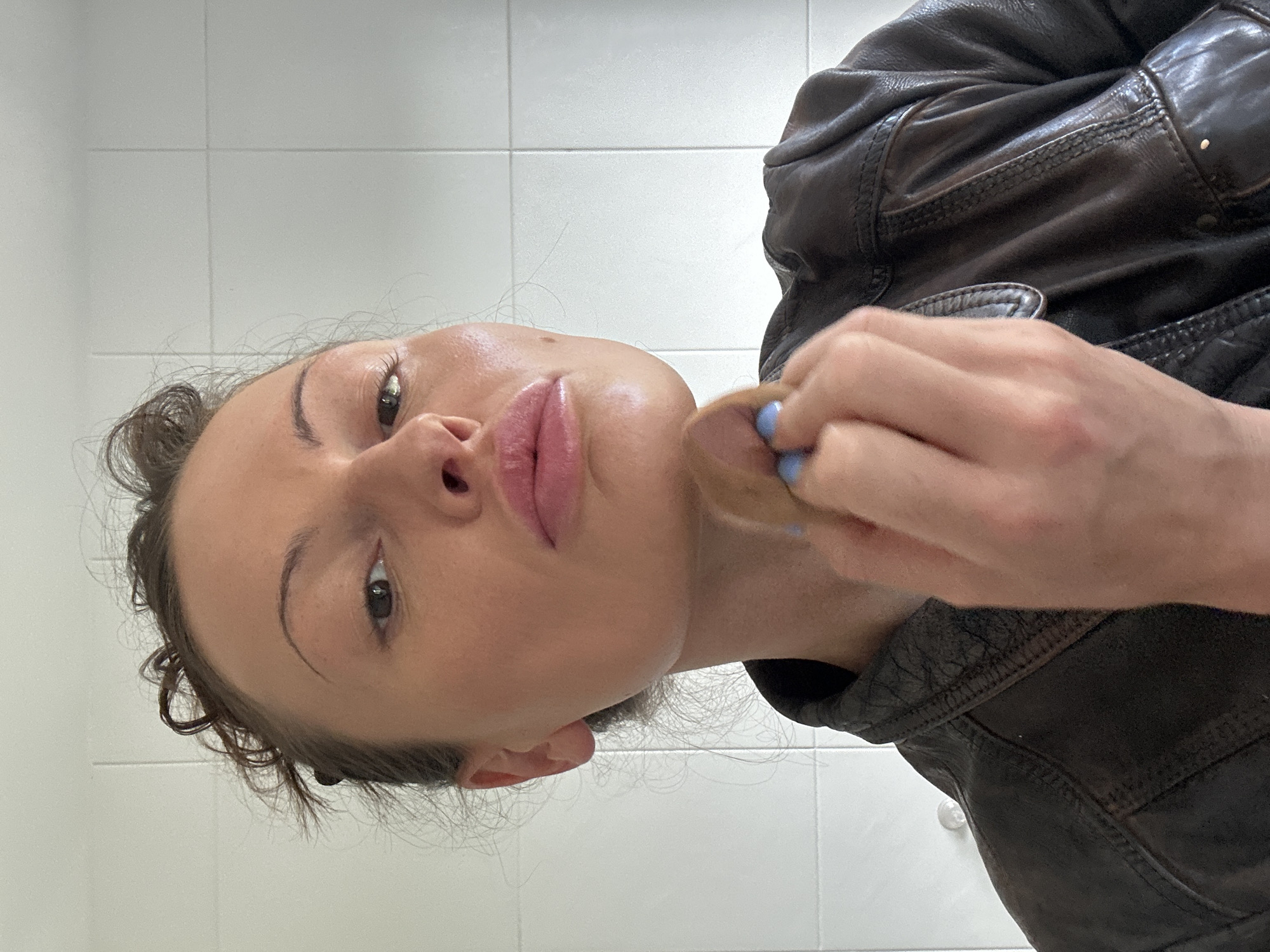 A woman applies makeup in a bathroom with white tiled walls. She holds a makeup sponge in her right hand, dabbing it near her face. She wears a dark brown leather jacket. The location is undetermined.
