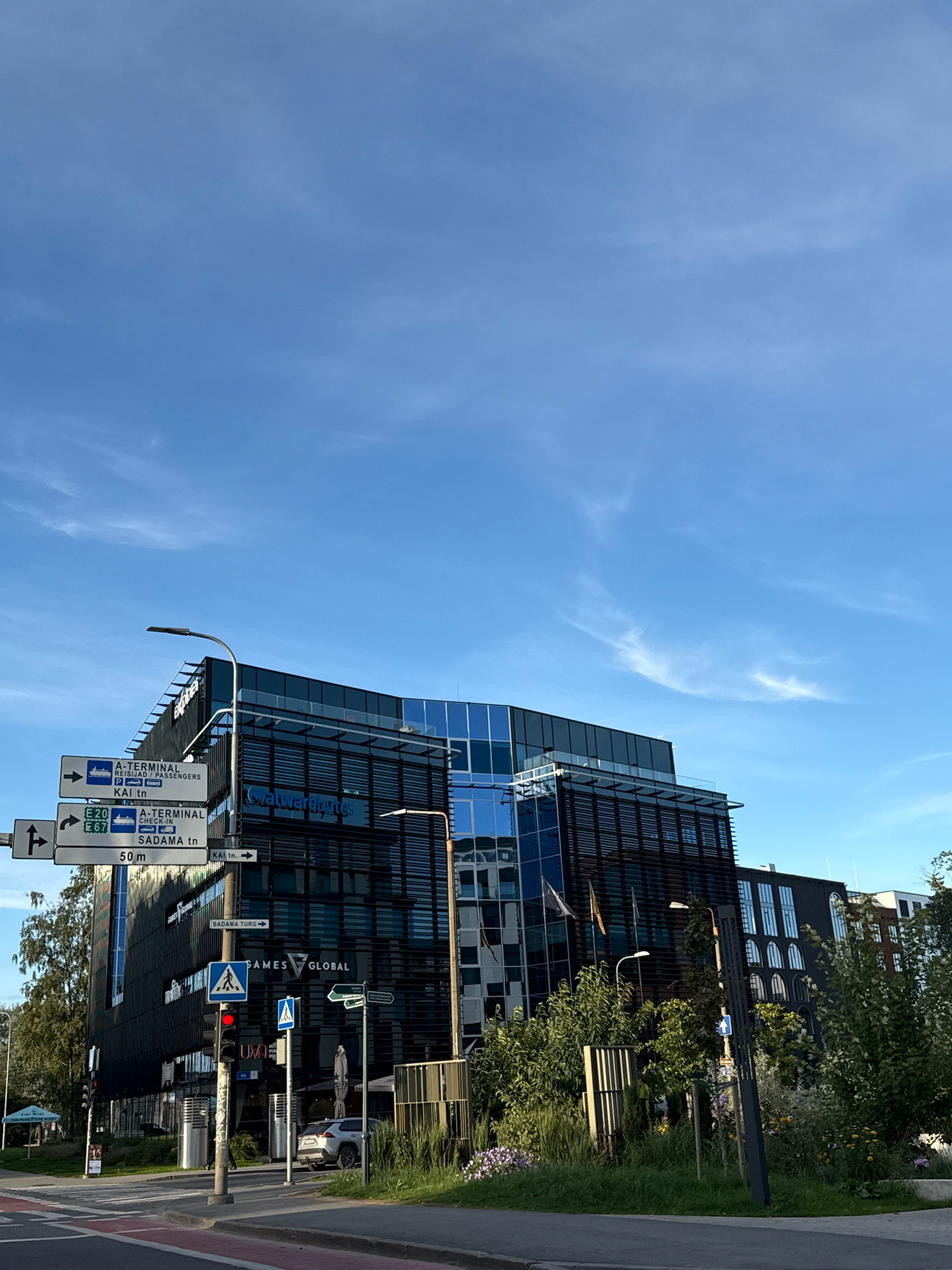 This image captures a view of the Rotermanni Quarter in Tallinn, Estonia, on a bright, clear day with a light blue sky and scattered wispy clouds. The focal point is a modern, multi-story building with a dark facade and large glass windows, housing the company "Matwartbytes" and "GAMES GLOBAL." Several directional signs are visible, indicating routes to the A-Terminal (for passengers and check-in), E20, E67, and "SADAMA tn." A traffic light displays a red signal, and a pedestrian crossing is marked on the road. Trees and greenery surround the building, and a white car is parked nearby. The scene appears to be an urban environment with architectural elements characteristic of contemporary city design.