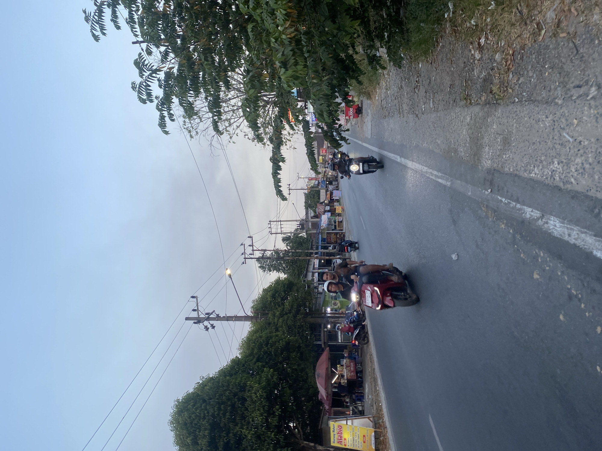 The image depicts a street scene in Banjarbaru, Indonesia, likely during dusk. The road is paved and marked with white lines. Several motorbikes are visible, some with riders. In the foreground, a red motorbike carries two men. Further down the road, another motorbike with a single rider is seen. On the left side of the road, there are small stalls or shops with umbrellas and signage, including one that reads "Alabio" and another with text in Indonesian. Trees line the left side of the road, and utility poles with wires are present overhead. The right side of the road features a concrete path and a patch of overgrown vegetation. The sky is overcast with a soft, diffused light.