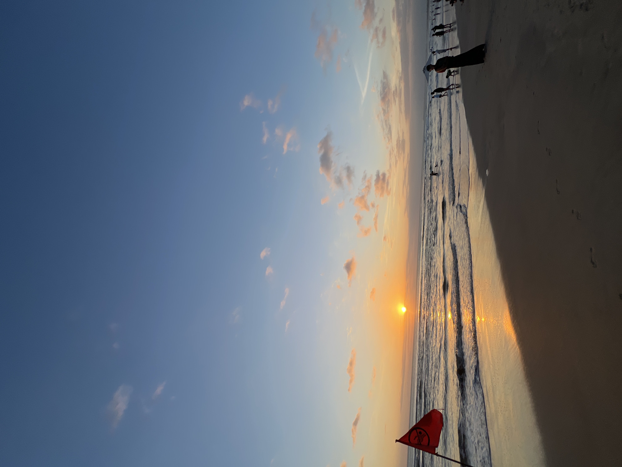This image captures a sunset scene at Kuta Beach in Kuta, Indonesia. The sun is low on the horizon, casting a warm orange glow across the sky and reflecting on the ocean waves. A red flag with a circular symbol is planted in the sand near the water's edge, indicating a warning. Several people are visible on the beach and in the shallow water, some walking along the shore, while others are wading or playing in the waves. A woman in a dark long dress stands on the sand, facing away from the camera, with a dog beside her. The sky is a gradient of blue and orange, with scattered clouds tinged pink by the setting sun. The wet sand on the beach shows footprints and is illuminated by the sunset's reflection.