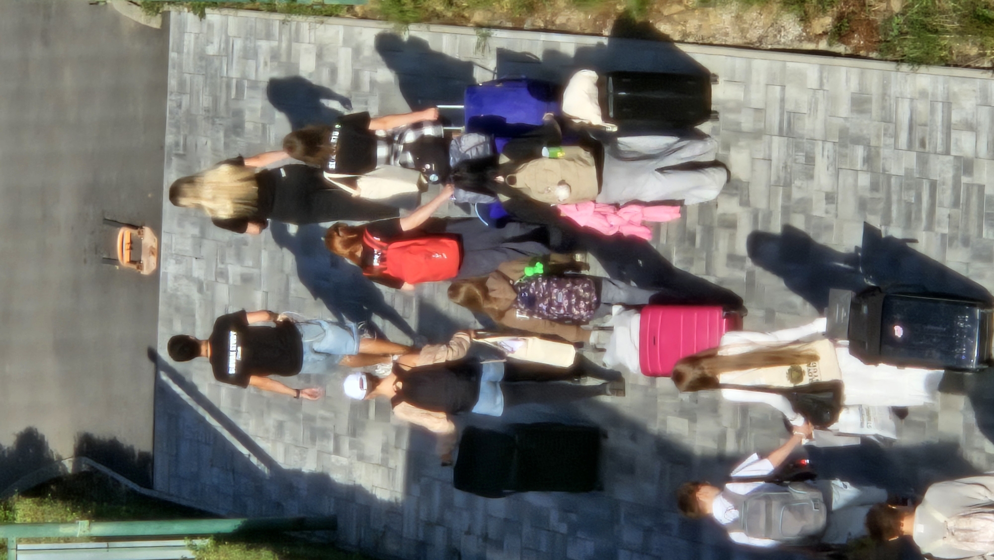 The image shows a group of individuals walking up a paved pathway in a city, possibly Prešov, Slovakia. The pathway appears to be made of interlocking grey stones. The individuals are carrying various pieces of luggage, including suitcases, backpacks, and bags.
In the foreground, a person with a grey backpack is walking, hand-in-hand with another person wearing a white dress and carrying a beige tote bag. To their right, a large pink suitcase is visible. Further back, a person in a brown hoodie with a patterned backpack walks alongside a person in grey pants with a beige backpack.
In the middle ground, a person in a tan top and denim shorts is walking with a black backpack and a black suitcase to their left. Next to them, a person in a red backpack walks with a black shirt. Another person in a black shirt and plaid skirt pulls a blue suitcase.
In the background, two individuals are walking away from the camera. One is wearing a black t-shirt with a graphic and light blue shorts, carrying a white rope. The other is wearing a black t-shirt and dark pants, with long blonde hair.
The environment appears to be outdoors, with some greenery visible on the right side of the image. Shadows are cast on the pathway, indicating daylight. A small orange object, possibly a piece of equipment or furniture, is visible on the pathway in the distance.