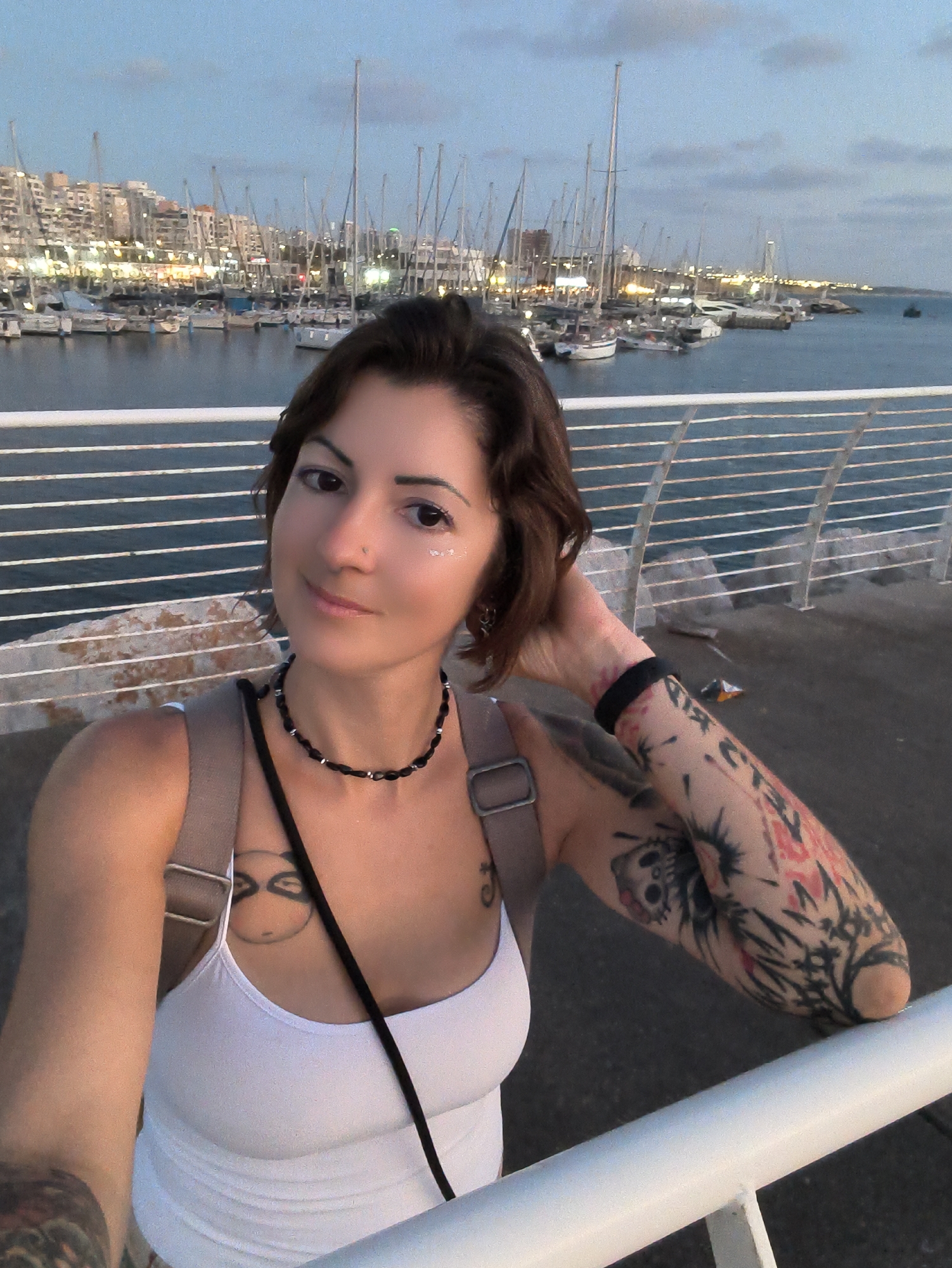 A woman with dark hair and tattoos on her arms is standing on a pier in the city of Ashkelon, Israel. She is wearing a white tank top and a black necklace. In the background, there is a harbor with many sailboats and buildings. The sky is blue and there are some clouds. The woman is looking at the camera and smiling. She has glitter on her cheek.