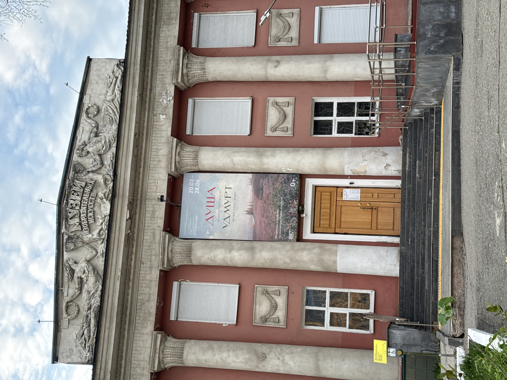 The image shows the facade of the Udmurt Republican Museum of Fine Arts in Izhevsk, Russia. The building has a classical architectural style with columns and decorative reliefs. A large banner advertising the "Soul of Udmurtia" exhibition is displayed between the columns. The entrance has stairs with a metal railing. A small sign is attached to the wall next to the stairs.