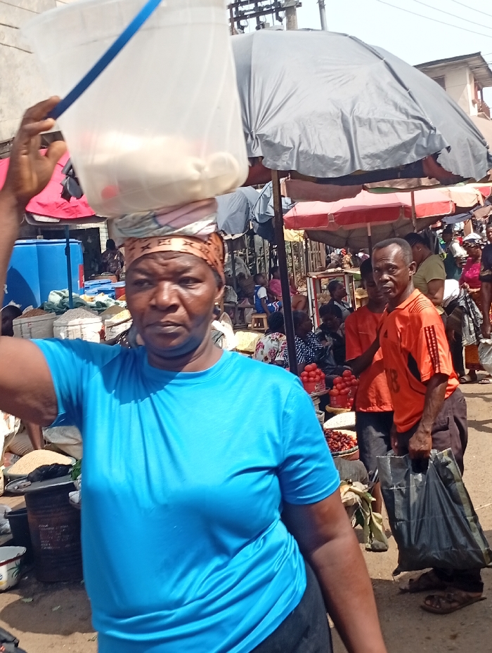 The image depicts a bustling outdoor market scene in Onitsha, Nigeria, characterized by numerous people, makeshift stalls, and merchandise under daylight conditions.
In the immediate foreground, an adult female with dark skin, wearing a bright blue short-sleeved t-shirt and dark bottoms, looks directly at the viewer. She is balancing a large, clear plastic bucket with a blue handle on her head, supported by a multi-patterned headwrap. Her right hand is raised, lightly touching the top edge of the bucket, which contains light-colored, amorphous contents, possibly food items, and a small red object.
Behind her, the market extends into the midground and background. An adult male with dark skin, wearing an orange short-sleeved shirt and dark trousers, stands or walks towards the right side of the frame, carrying a large black plastic shopping bag in his left hand. His gaze is directed to his right. Adjacent to him, a stall displays numerous red tomatoes in clear containers, along with other produce.
The environment is densely populated with other market-goers and vendors, some seated, some standing, under various sunshade structures. A large, grey canopy umbrella dominates the upper-midground, with smaller red and other colored canopies visible. Sacks of goods, likely grains or produce, are visible near the stalls. Overhead, power lines are present. The ground appears to be unpaved earth. The overall scene suggests a busy commercial thoroughfare within the city.