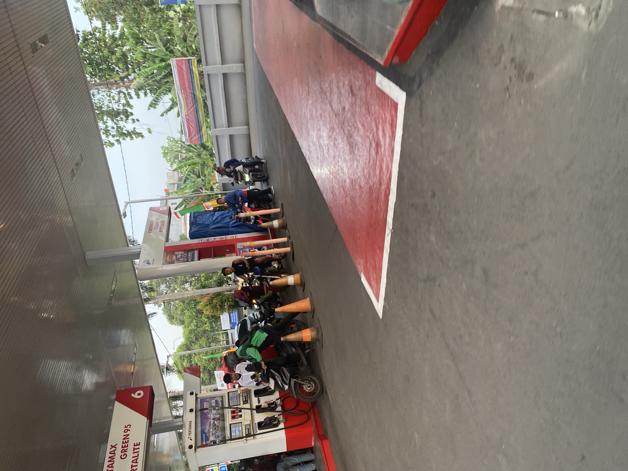 The image shows a Pertamina gas station in Depok, Indonesia. Several people are present, some on motorcycles, at the fuel pumps. One person in a green jacket is pushing a motorcycle at a pump. There are traffic cones on the ground. In the background, there are trees and a wall with banners. The overall scene depicts a typical day at a gas station.