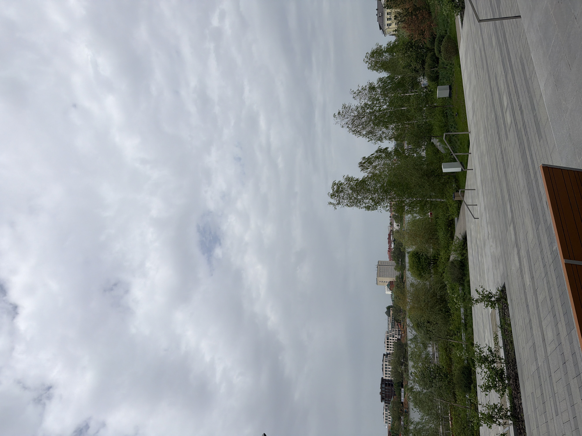 The image depicts a cloudy sky over a cityscape, likely taken in Kazan, Russia, based on architectural clues and the presence of birch trees which are common in the region. In the foreground is a modern paved area constructed from light grey brick. A low retaining wall of the same material runs parallel to a line of trees. This wall has vertical metal bars protruding from the top, as well as boxy white lights. Behind the trees is a park-like area with dense vegetation and some exposed soil. In the background, buildings of varying heights are visible, mostly white or light-colored, suggesting an urban environment. The sky is predominantly overcast, and light and color are diffused.