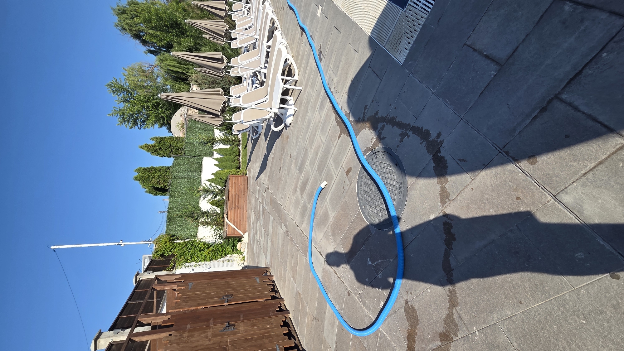 A blue corrugated hose lies on a paved area with a shadow of a person on the left. The hose curves from the left side of the frame towards the right, forming a large loop. There is a circular manhole cover in the center of the frame with text that reads "RECO" and "ISO 9001-2000". In the background, there is a wooden fence, a row of lounge chairs with closed umbrellas, and lush green trees. The sky is clear and blue. The scene appears to be outdoors in a sunny environment, possibly near a pool area.