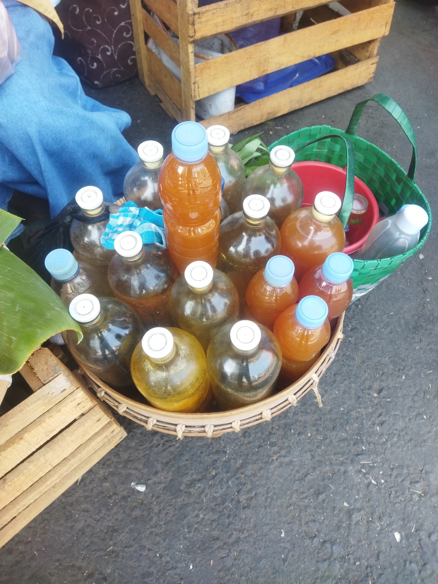 In the city of Wonogiri, Indonesia, a close-up, high-angle shot captures a collection of glass bottles filled with amber-colored liquid, arranged in a woven basket. The bottles vary in size and shape, with some featuring white caps and others blue. A large, clear plastic bottle with a white cap is also present. A green woven bag is partially visible in the background, containing a red bucket. To the left, a piece of a large green leaf and a wooden crate with slatted sides are present. The bottles appear to be for sale, suggesting a market or roadside stall setting. The ground beneath is asphalt, with some debris scattered on its surface.