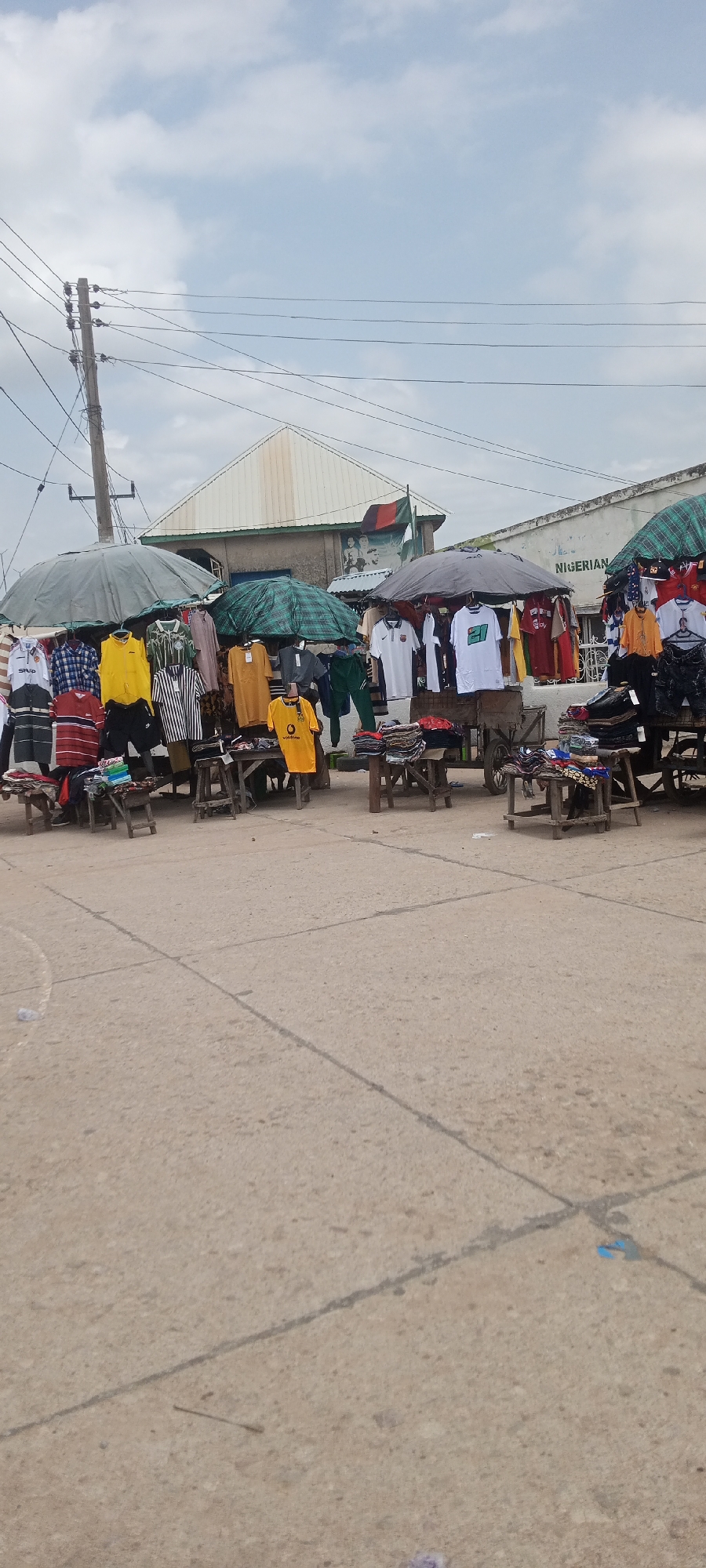 The image depicts an outdoor clothing stall in Jalingo, Nigeria. The stall is set up on a paved surface, likely a street or market area, with a cloudy sky overhead. Several racks of t-shirts and other apparel are displayed under large umbrellas, suggesting a sale or market day. The clothing items appear to be predominantly t-shirts, with various colors and designs visible, including some with sports team logos or branding. Some garments are neatly folded on wooden tables, while others hang from racks. In the background, there are buildings with a distinctive roof structure and a sign that reads "NIGERIAN." The scene suggests a bustling commercial environment, common in Nigerian cities.