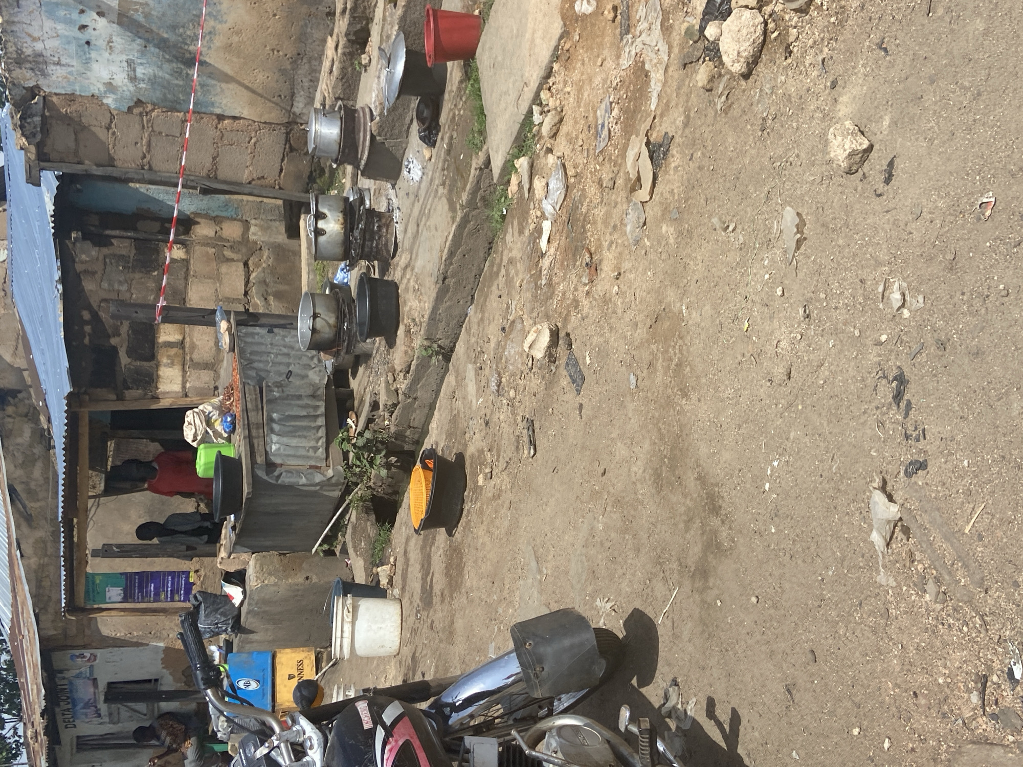 This media file depicts an outdoor scene in Bukuru, Nigeria. The setting appears to be a marketplace or a roadside stall area with a rustic, informal construction. A building with brick walls and a corrugated metal roof is visible. In front of this structure, several metal pots, likely for cooking, are stacked and arranged. Some appear to be in use or recently used, with soot stains visible. A red bucket is placed on top of one of the pots.
The ground is a mix of dirt, sand, and small stones, with scattered debris, suggesting an unpaved area. A concrete pathway or ledge runs through the scene. Several plastic containers, including white buckets and a green basin, are also present.
In the background, through an opening in the structure, figures of people can be seen, some wearing red and gray clothing. One person is partially obscured by a green basin and a black bowl. The presence of these individuals suggests that this is an active location, likely a place of business or daily life.
Further to the right, a motorcycle is partially visible, with its handlebars and front fender in the foreground, indicating a transportation element within the scene. An orange safety reflector is lying on the ground near the concrete.
The lighting suggests it is daytime, with shadows visible. The overall atmosphere is one of everyday commerce and activity in a somewhat informal environment. There is some text visible on posters and crates in the background, but it is largely illegible due to image quality and distance.