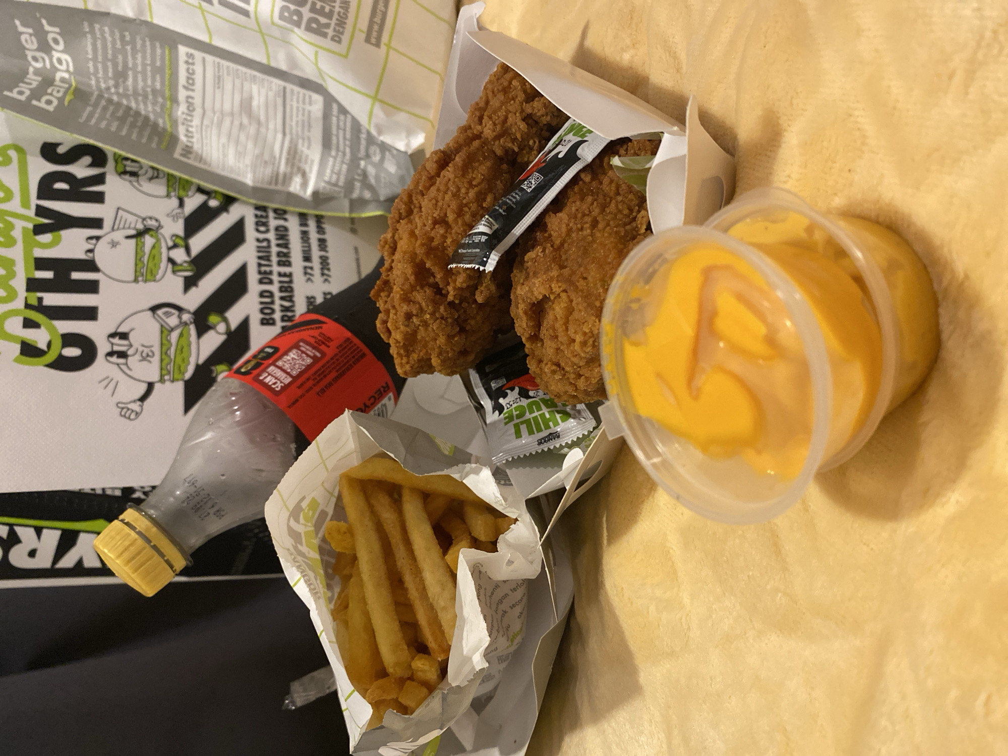 The image depicts a fast-food meal seemingly from "Burger Bangor," a restaurant chain. The meal includes fried chicken, french fries, and a yellow sauce, possibly cheese or mayonnaise, in clear plastic containers. A bottle, possibly a beverage, also features in the image. Text visible indicates "72 Million Burgers" and "7200 Job Opportunities." The location is suggested to be Nusa Dua, Indonesia, from the "www.burgerbangor.com" URL and "DENGAN" text, indicating Indonesian language. No people or specific actions are visible in the frame. The food and packaging are casually arranged on a soft, tan-colored surface, likely a sofa or blanket. The image quality suggests an amateur photograph.