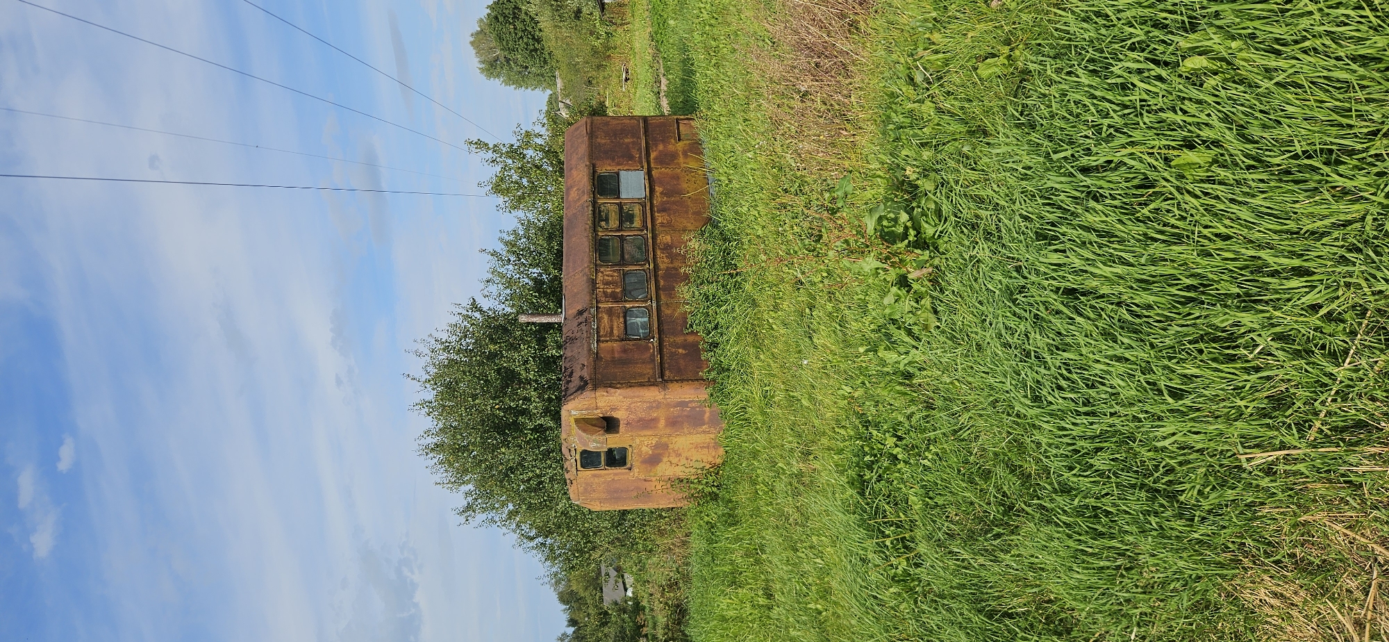 An old, rusted caravan is parked in a grassy field with trees and power lines in the background. The caravan is oblong with several windows along one side and a chimney pipe protruding from the roof. The grass is tall and green, with some dry patches. The sky is blue with scattered clouds. The scene appears to be in a rural area.