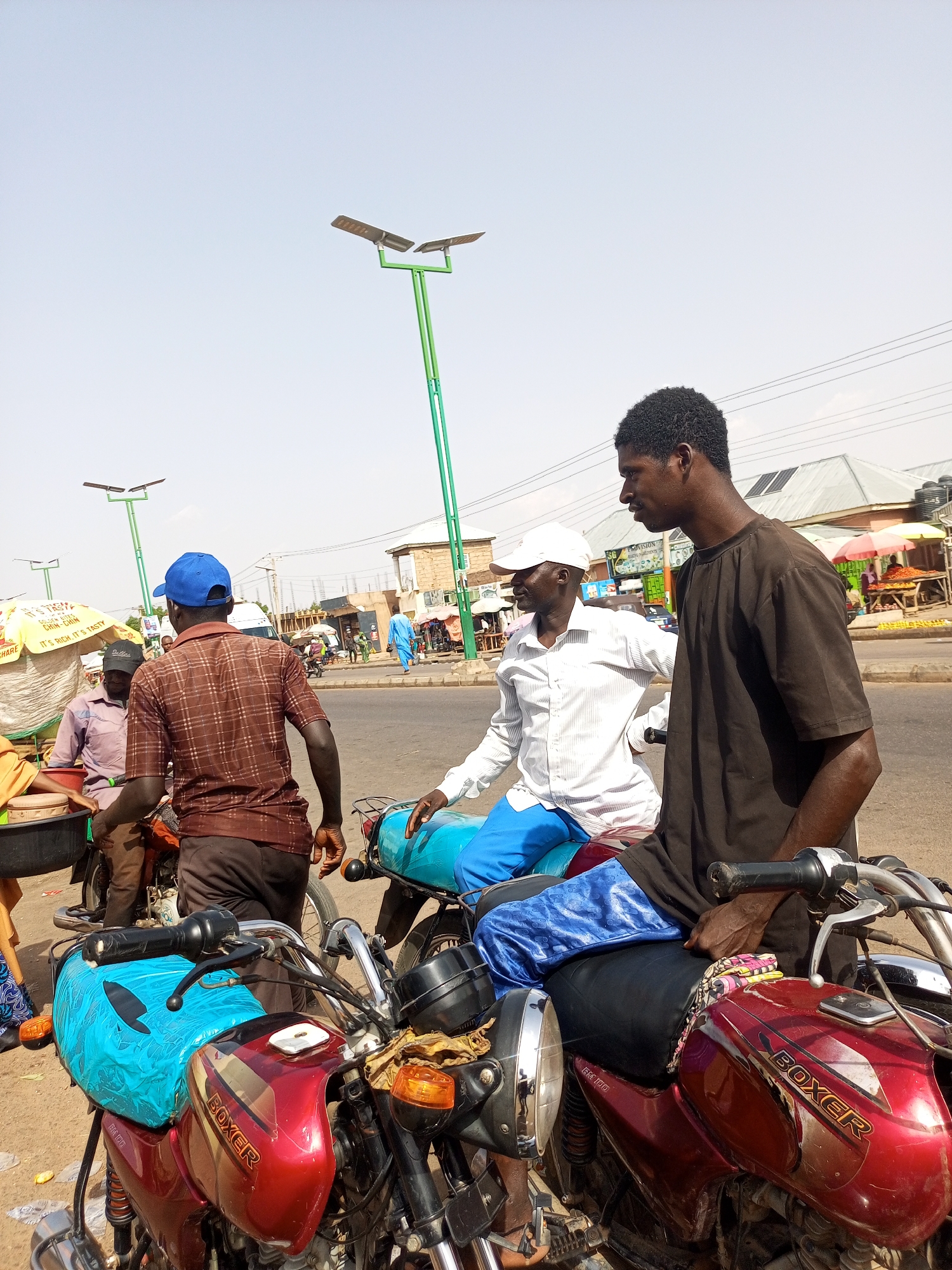 The image depicts a street scene in what appears to be Tempure, Nigeria. In the foreground, three men stand near motorcycles. Two of the motorcycles are red "Boxer" models. One man wears a brown t-shirt, another a white shirt and blue pants, and a third a brown checkered shirt and blue cap. Other people and vehicles are visible in the background, along with buildings, solar-powered streetlights, and roadside vendors with umbrellas. One umbrella has the text "IT'S RICH. IT'S TASTY" on it.