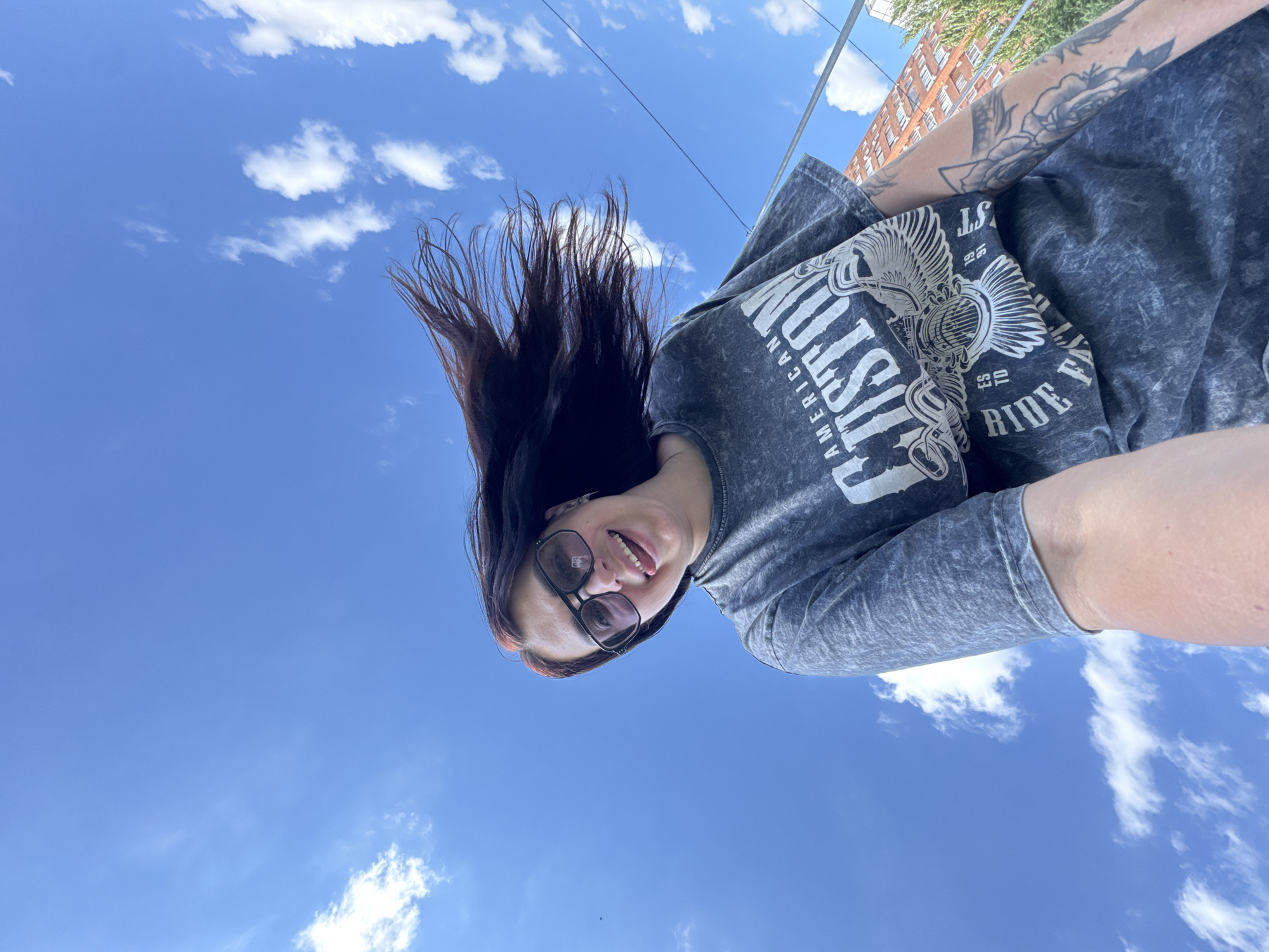 A low-angle, wide shot captures a young woman with long, dark hair blowing in the wind against a bright blue sky with scattered white clouds. She wears large, dark-rimmed sunglasses and a grey t-shirt with a graphic print that reads "AMERICAN CUSTOM" and "RIDE FAST". Her tongue is sticking out slightly to the left side of her mouth, and she appears to be looking up at the camera. Her right arm, visible on the right side of the frame, displays a tattoo of a bird and floral motifs. In the background, a multi-story brick building and green trees are visible, along with a utility pole and wires, suggesting an urban environment in Tolyatti, Russia. The perspective implies the photo was taken from a low vantage point, possibly from the ground or a low-lying object, looking upwards.