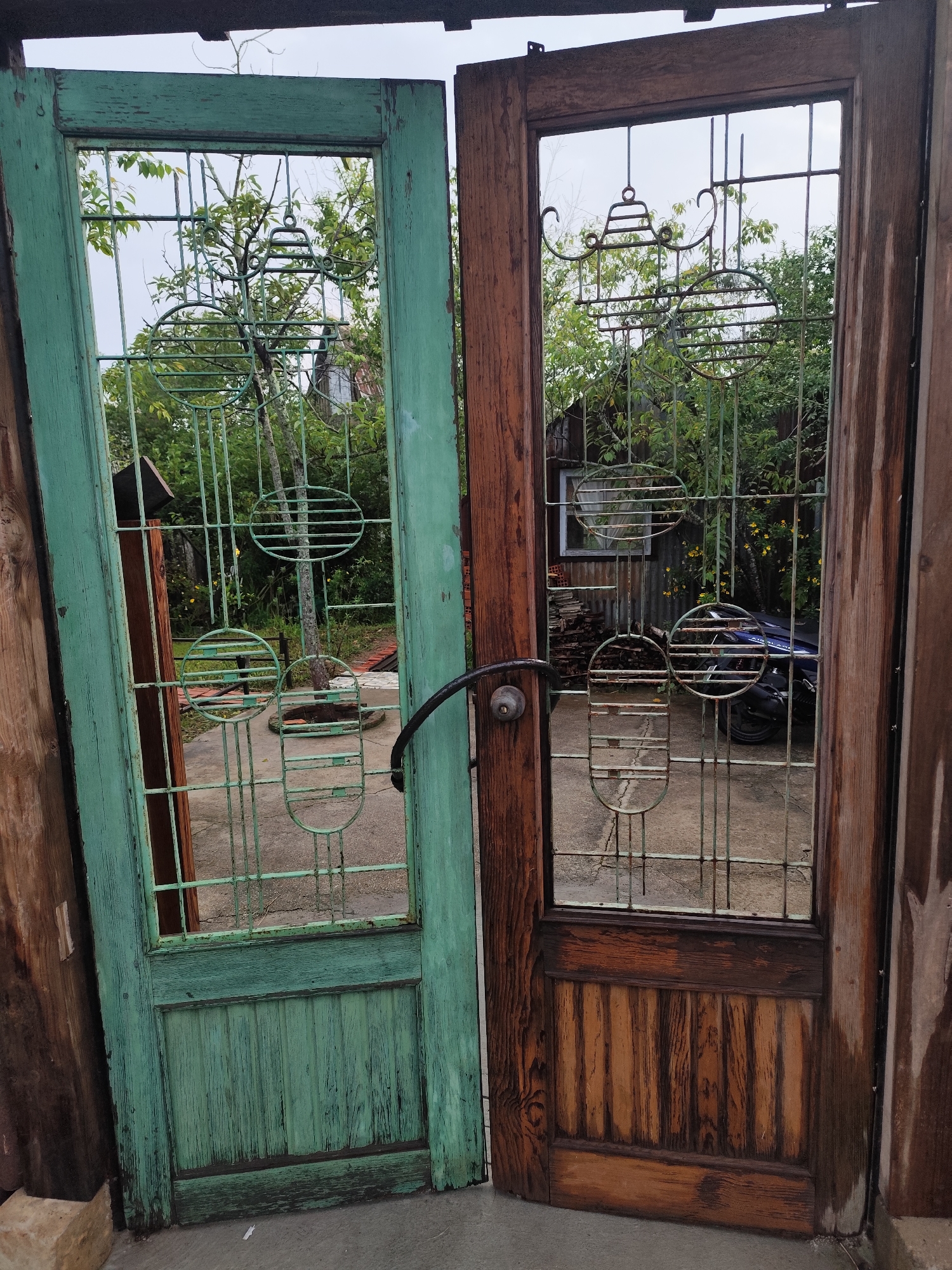 The image displays two old wooden doors, one painted green and the other unfinished wood, standing open side-by-side. Both doors feature decorative metal grilles in their upper sections, partially obscuring the view of a backyard. The green door has a weathered, peeling paint finish, while the wooden door shows prominent grain patterns. A black metal door handle is attached to the right-side door. Beyond the doors, a blurred background reveals trees, foliage, a rustic building with a window, a stack of bricks, and a dark-colored motorcycle. The ground appears to be concrete. The setting suggests a residential area, possibly a yard or an alleyway.