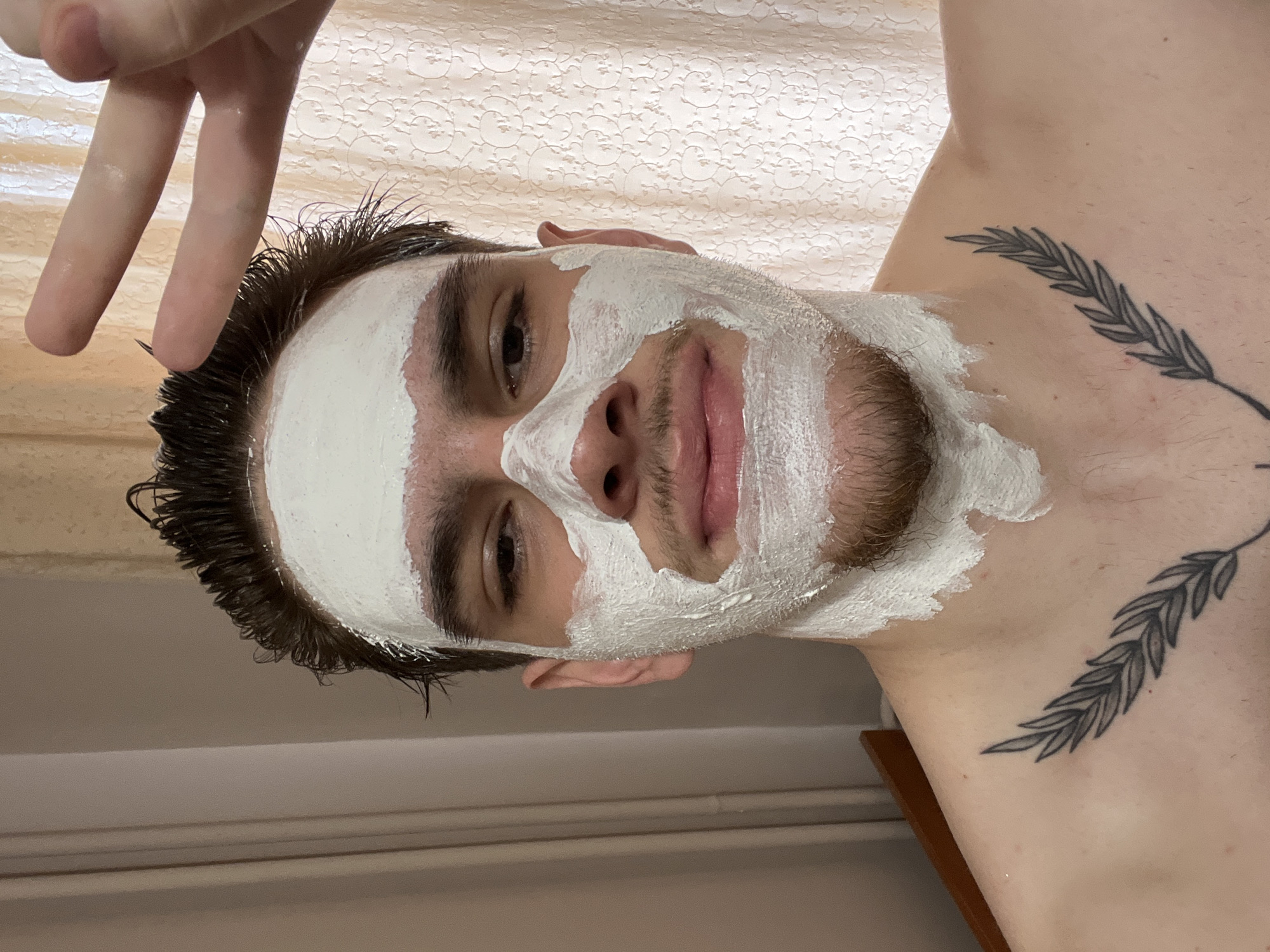 A man is depicted in a close-up, tilted head and shoulders shot. He has a white facial mask applied to most of his face, excluding areas around his eyes, lips, and beard. His dark hair is wet and slicked back. He is making a peace sign with his left hand near his forehead. On his upper chest, there is a tattoo of a leafy branch. The background shows a wall with a textured, off-white patterned surface. The location in Sofia, Bulgaria, is unknown based on the visual information provided.