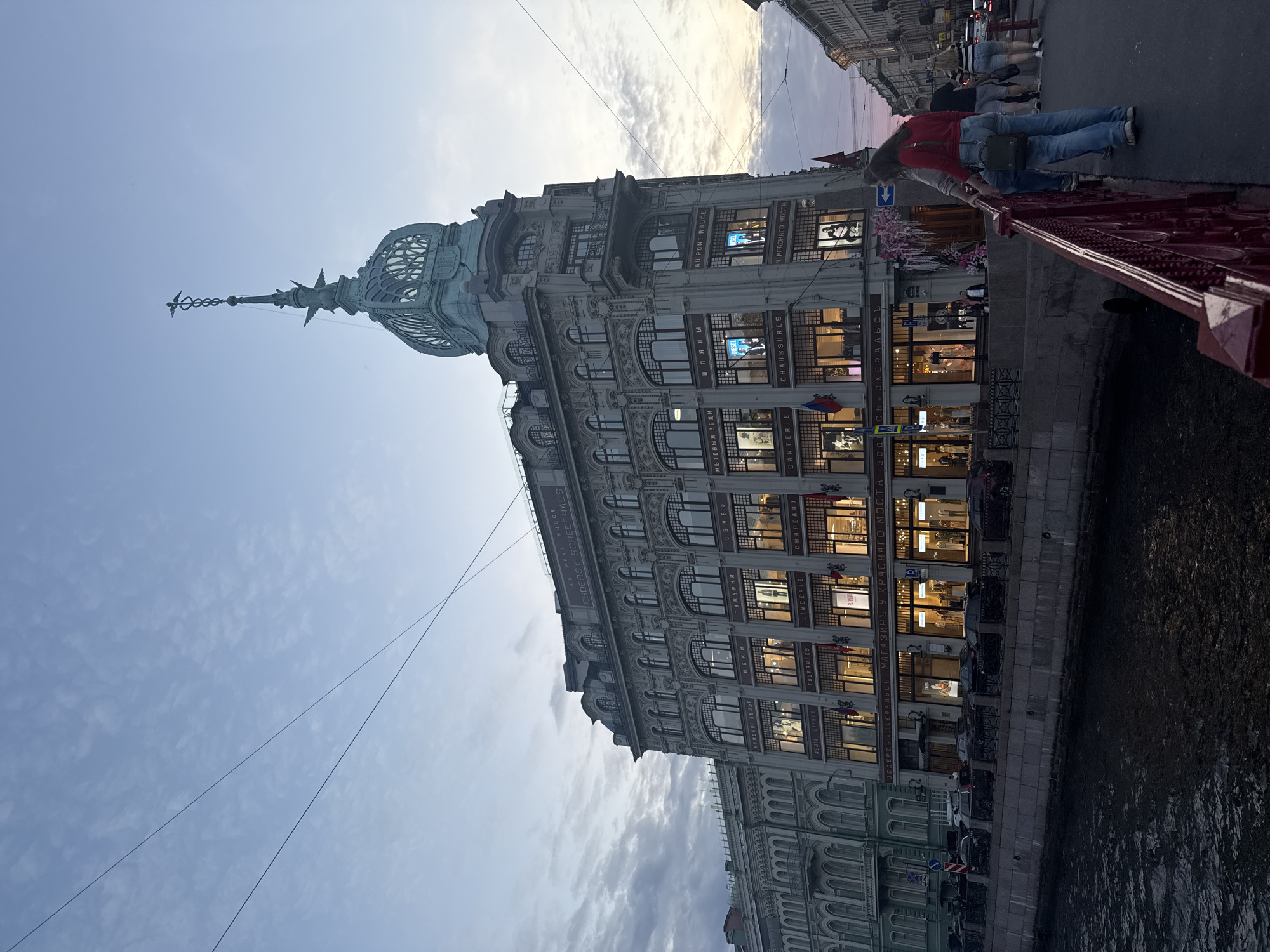 This image captures a vibrant urban scene in Saint Petersburg, Russia, featuring the historic Esders and Scheefhals building, now known as DLT (Dom Leningradskoy Torgovli), alongside a canal. The grand, multi-story building dominates the left side of the frame, showcasing ornate architecture, numerous large windows with warm interior lighting, and a distinctive dome topped with a spired finial resembling a caduceus.
In the foreground on the right, a decorative red metal railing of a bridge extends into the composition, framing a section of the dark canal water. Several people are visible on the bridge; one individual wearing a red top and blue jeans is leaning against the railing, looking towards the canal and building. Others are standing further back on the bridge.
The sky above displays a mix of light blue and scattered clouds, suggesting either late afternoon or early evening, a time when artificial lights inside buildings become noticeable. Power lines crisscross the upper sky.
Visible text on the building's facade provides strong location and identity cues. "ESDERS ET SCHEEFHALS" identifies the historic department store. "AU PONT ROUGE" (At the Red Bridge) and its Russian equivalent "У КРАСНАГО МОСТА" are seen, referring to the Red Bridge in the foreground. Department names such as "МУЖСКОЕ БЫЛЬЕ" (Men's Underwear), "шляпы" (hats), "CHAUSSURES" (shoes), "GANTÉRIE" (gloves), "ЛИНЖЕРИ" (lingerie), and "ФУРНИТУРА" (haberdashery) are also discernible. The scene presents a blend of historical architecture and modern city life.