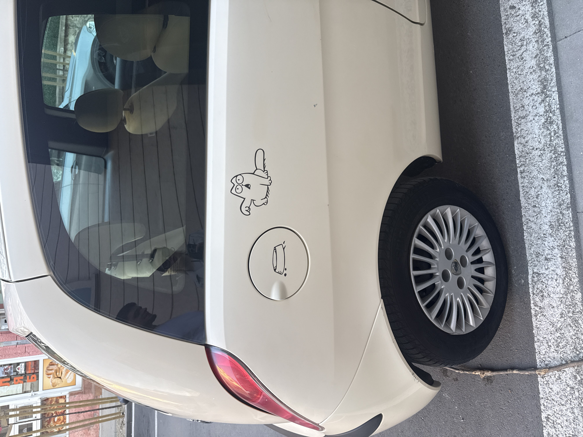 The image shows the rear quarter of a white car parked on a street. The car has a cartoon cat sticker and a fuel filler door with a tire inflation symbol. A portion of the rear passenger window is visible, reflecting the interior of another car. The rear tire of the car is visible. To the left of the car, a storefront with posters depicting bakery items can be seen. The car is parked on a street with a white line marking the edge of the lane. Based on the storefront signage in the background, the location is in Blagoevgrad, Bulgaria.