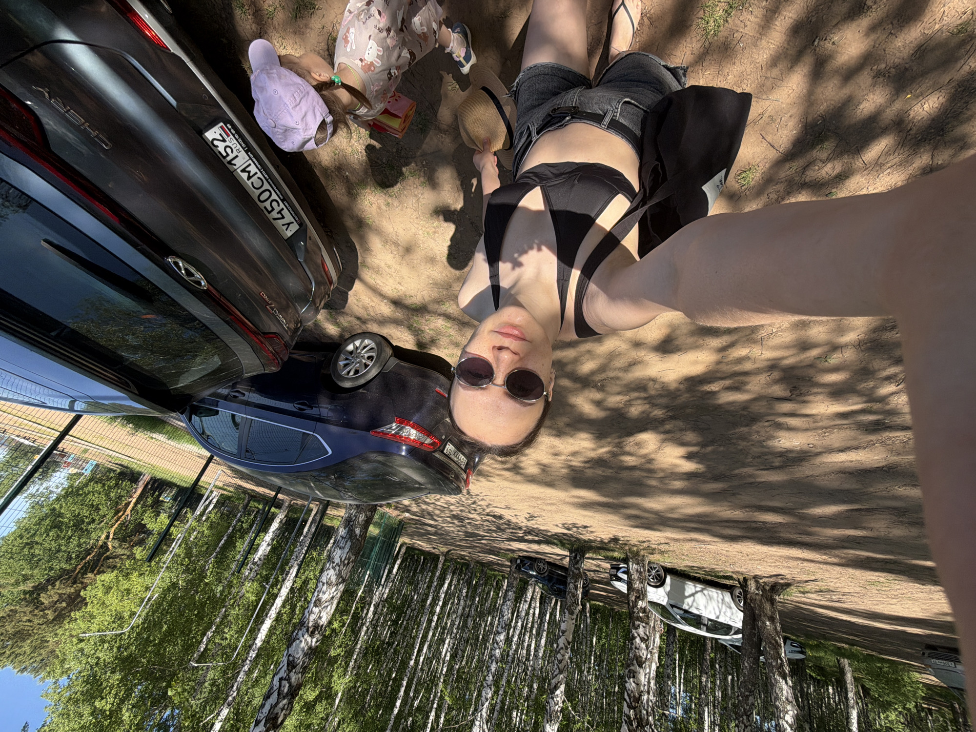 The image is taken from a high-angle perspective with the camera rotated 180 degrees. It appears to be a selfie taken outdoors, possibly near a wooded area in or near the city of Chaikovsky, Russia.
In the foreground is a woman wearing sunglasses and a black bikini-style top with black denim shorts. Her right arm is extended towards the camera. Next to her is a child wearing a pink patterned outfit and a lavender hat. Beside the woman is a light-colored straw hat.
Two cars are parked nearby. The closest car is dark gray, identified as a Chery Tiggo 7 Pro based on the badging. Its license plate reads "Y450CM 152 RUS." A second car, a dark blue sedan, is also parked next to the Chery.
The background consists of a dense area of birch trees with white trunks, suggesting a forest or park setting. Sunlight filters through the trees, casting shadows on the ground. Two more cars are visible among the trees.
