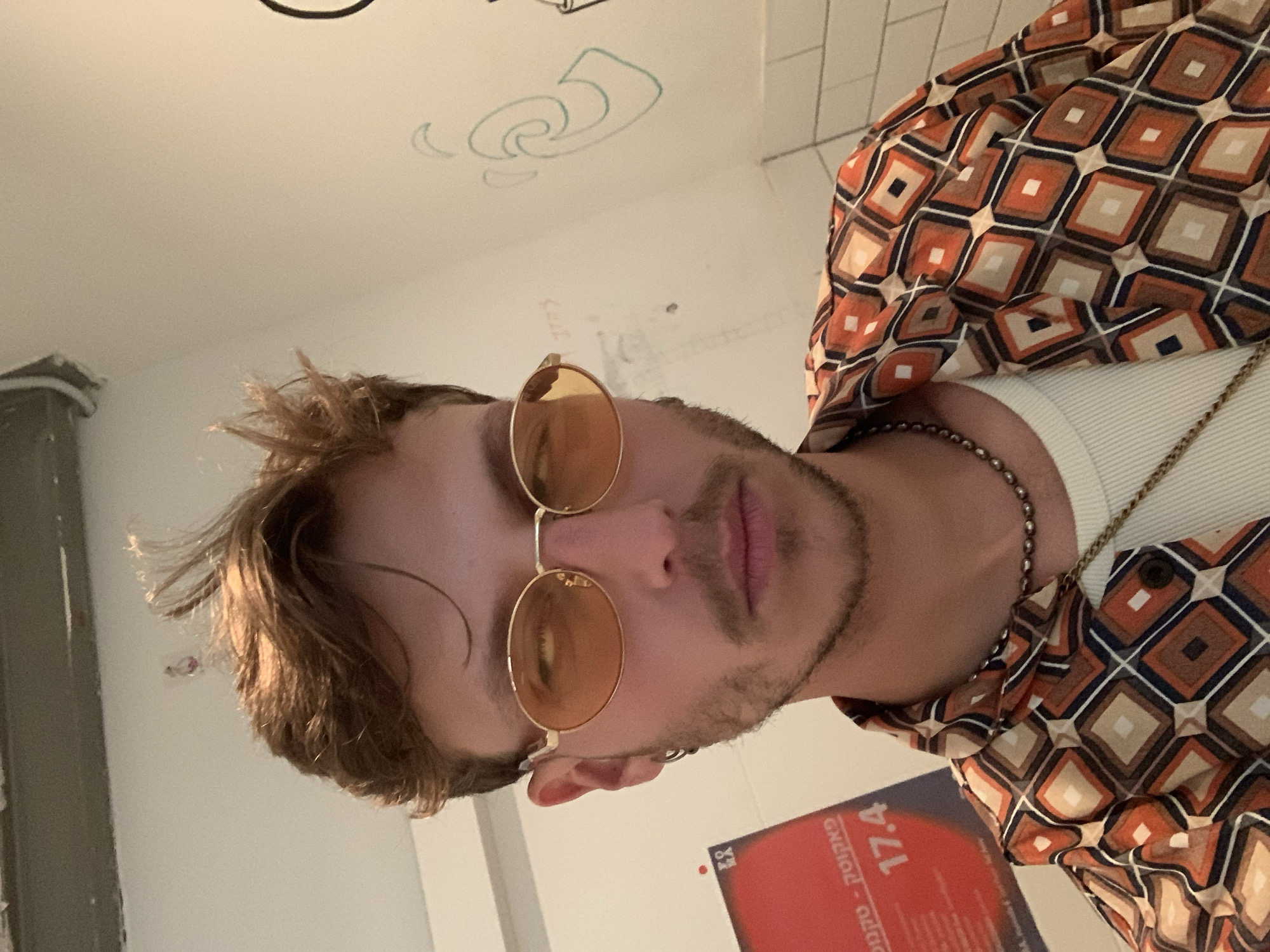 A portrait of a man with light brown hair and a trimmed beard, wearing round-frame sunglasses, a patterned shirt with orange, brown, and white squares, and two necklaces (one beaded, one a thin gold chain). He is partially visible, facing the camera at an angle. He is standing in a bathroom. The wall behind him is white with some doodles. There is a red object partially visible at the bottom, partially covered by his clothing. The red object has the word "פאקס" written on it.
The city of Tel Aviv, Occupied Palestine is inferred only from the text appearing on the object in the picture.