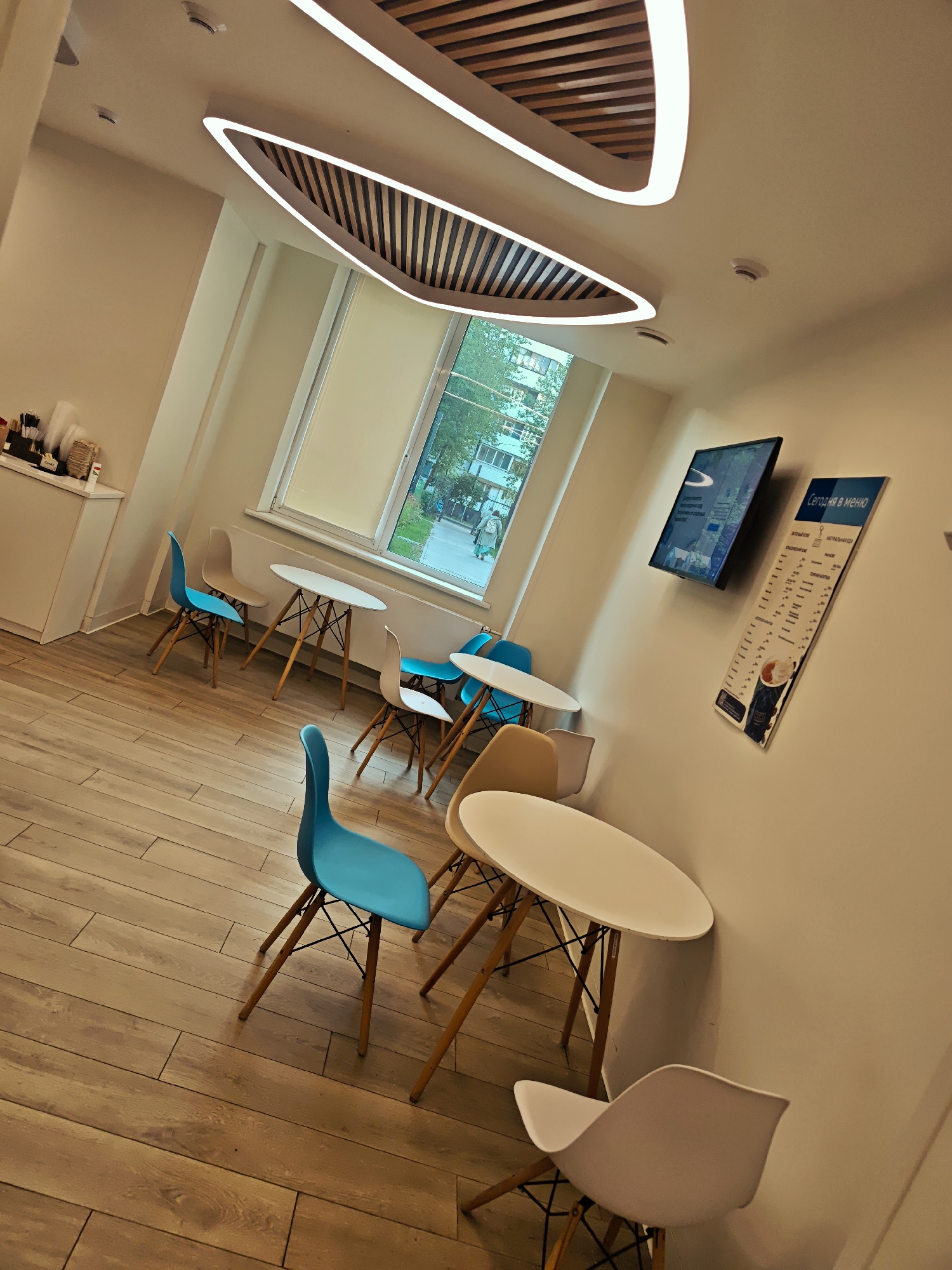 This image captures an interior view of a modern and brightly lit cafe or break room, likely in Moscow, Russia, given the context. The space is characterized by a clean, minimalist design with a warm color palette.
The main subjects are the furniture and architectural elements. There are several small round white tables with wooden, splayed legs and black wire supports, each accompanied by two to three chairs. The chairs are Eames-style, featuring plastic bucket seats in a mix of vibrant turquoise/light blue, off-white, and light tan/beige, also with wooden legs and black wire supports. The floor is covered in light-colored wooden planks laid in a staggered pattern.
The ceiling features a distinctive, large, oval-shaped light fixture with an illuminated LED strip outlining its wavy contours. Inside this fixture, and extending along the ceiling, are parallel wooden slats, adding a natural and decorative touch. Two small smoke detectors or sensors are visible on the plain white ceiling.
On the left, a white counter or cabinet with a dispenser for cups and other items is partially visible. A large window dominates the back wall, offering a glimpse of the outdoors – green trees and multi-story apartment buildings, suggesting an urban setting. The lighting suggests it is daytime.
On the right wall, there is a wall-mounted flat-screen television displaying what appears to be digital content or advertisements. Next to it, a large vertical menu board is affixed, titled in Russian: "Сегодня в меню" (Today on the menu). Below the title, a list of food and drink items is visible, including categories like "КЛАССИКА" (Classics) and "НАТУРАЛЬНАЯ ЕДА" (Natural Food), along with various beverages like coffee, tea, and other options.
There are no people visible in the image, implying the space is currently unoccupied, perhaps awaiting customers or captured during off-peak hours. The overall impression is one of a contemporary, inviting, and functional communal space.