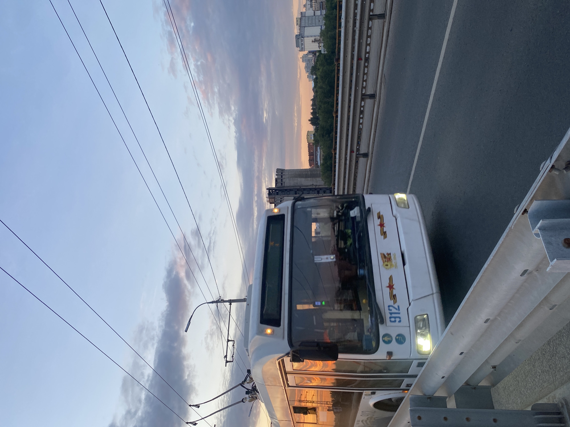 A white trolleybus with the number 912 is traveling on a bridge in Novokuybyshevsk, Russia. The trolleybus is in the foreground on the right side of the image, with its headlights on. The sky above is a mix of blue and grey clouds, with streaks of orange and pink from the sunset. In the background, there are several buildings, including a tall, grey cylindrical structure and other modern buildings. Trees line the river or bank below the bridge. The bridge has a metal guardrail on the right side. The overall scene captures a moment of urban transportation at dusk.