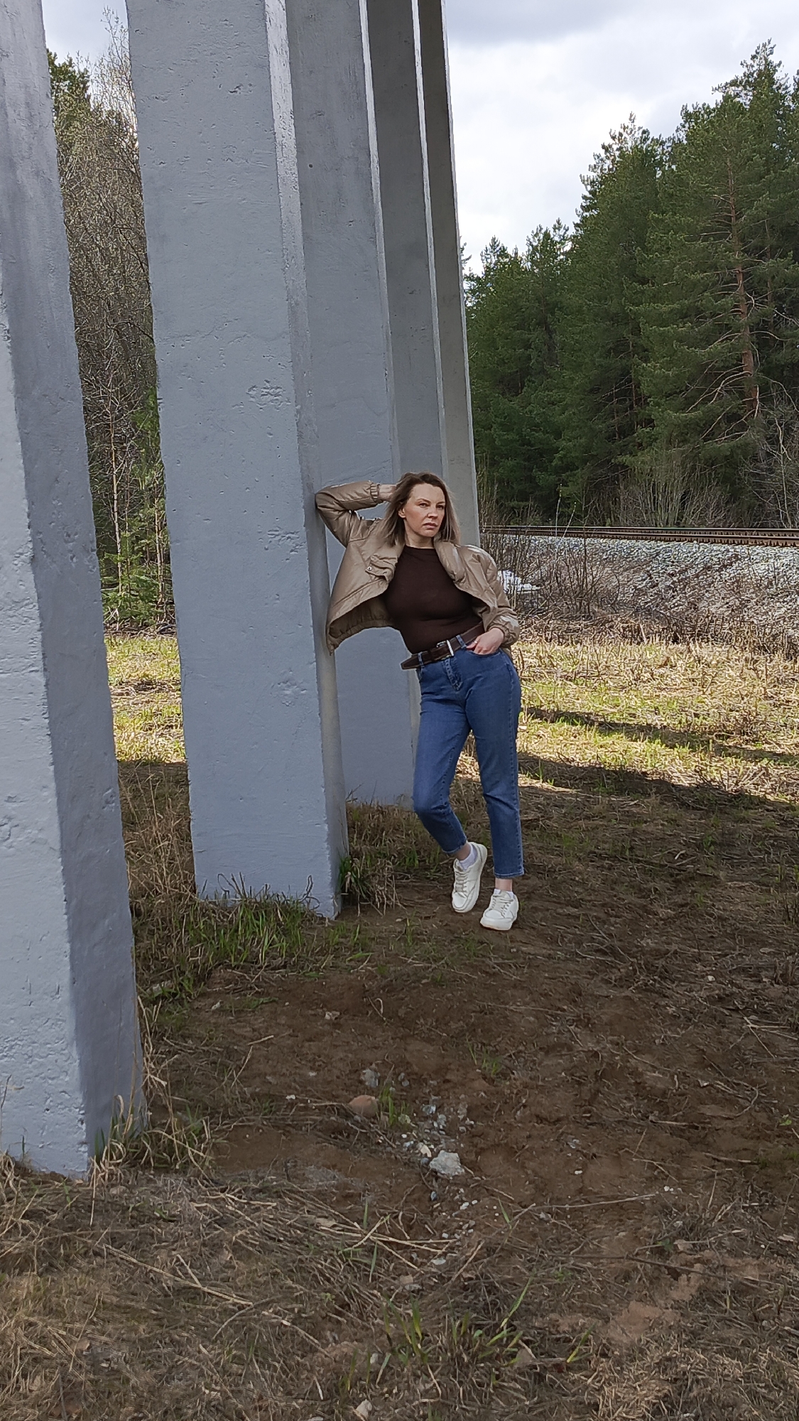 A woman poses outdoors, leaning casually against a large, light grey concrete pillar. She wears a light brown bomber jacket over a dark brown long-sleeved top, blue jeans, and white sneakers. Her light brown hair falls around her shoulders, and she looks to the right with a calm, composed expression, one hand resting on her head/hair and the other in her jeans pocket.
The scene depicts a blend of infrastructure and nature, likely in Chaikovsky, Russia. Several similar concrete pillars extend to her left, suggesting a bridge, overpass, or industrial structure. The ground beneath her is uneven, a mix of dry, light brown grass, sparse patches of green vegetation, and exposed dirt with small stones. In the background, a dense forest of tall pine trees creates a natural backdrop. Faintly visible beyond the trees on the right are railroad tracks. The sky is bright and appears partly cloudy or overcast, indicating daytime conditions with diffused natural light. There is no visible text in the image.