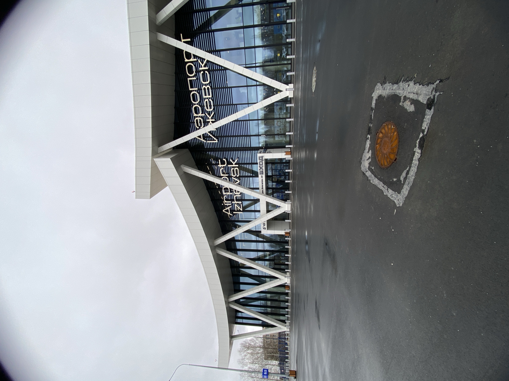 The image shows the Izhevsk Airport terminal in Russia. The facade features white structural supports with large glass windows. Signs in both Russian and English ("Аэропорт Ижевск" and "Airport Izhevsk") are visible above the entrance. The foreground is an asphalt parking area with a manhole cover. The sky is overcast.