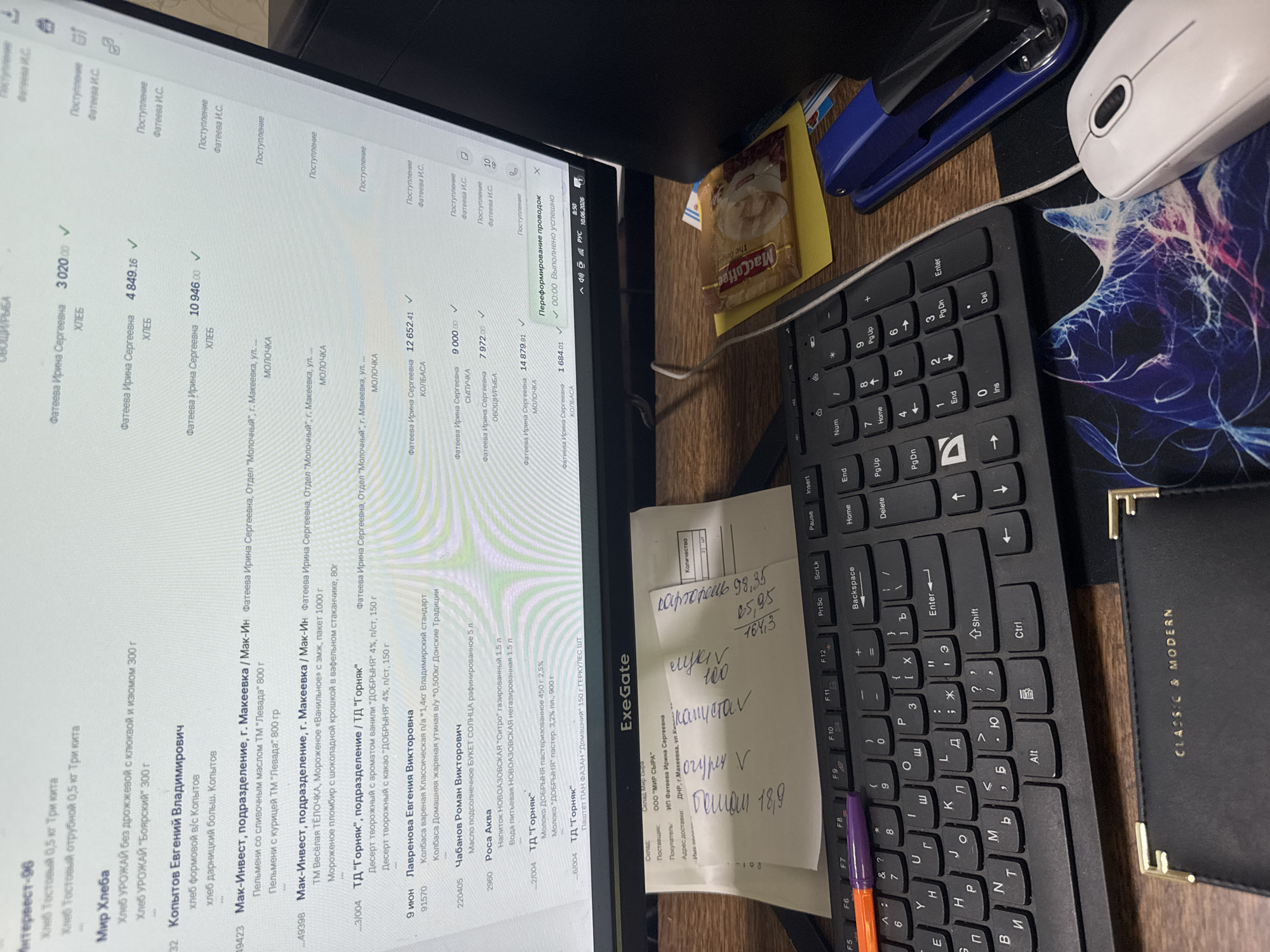 The image shows a computer screen displaying a list of items, likely an inventory or order system. The screen is positioned on a desk in front of a keyboard and a mouse. Several handwritten notes and office supplies, including pens and a stapler, are also visible on the desk. One of the notes is placed on top of a coffee packet. The background shows a monitor with a timestamp "10.06.2026" and "8:59". The overall scene suggests an office or workspace environment.