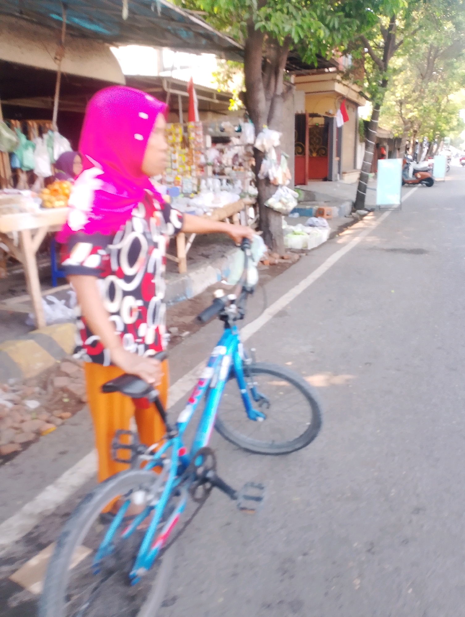 A woman wearing a bright pink hijab and a patterned shirt is standing on a sidewalk next to a blue bicycle. She is holding the handlebars of the bicycle. The background shows a street scene with shops, trees, and other bicycles. The image appears to be taken in Probolinggo, Indonesia, judging by the architecture and the presence of the Indonesian flag in the background.