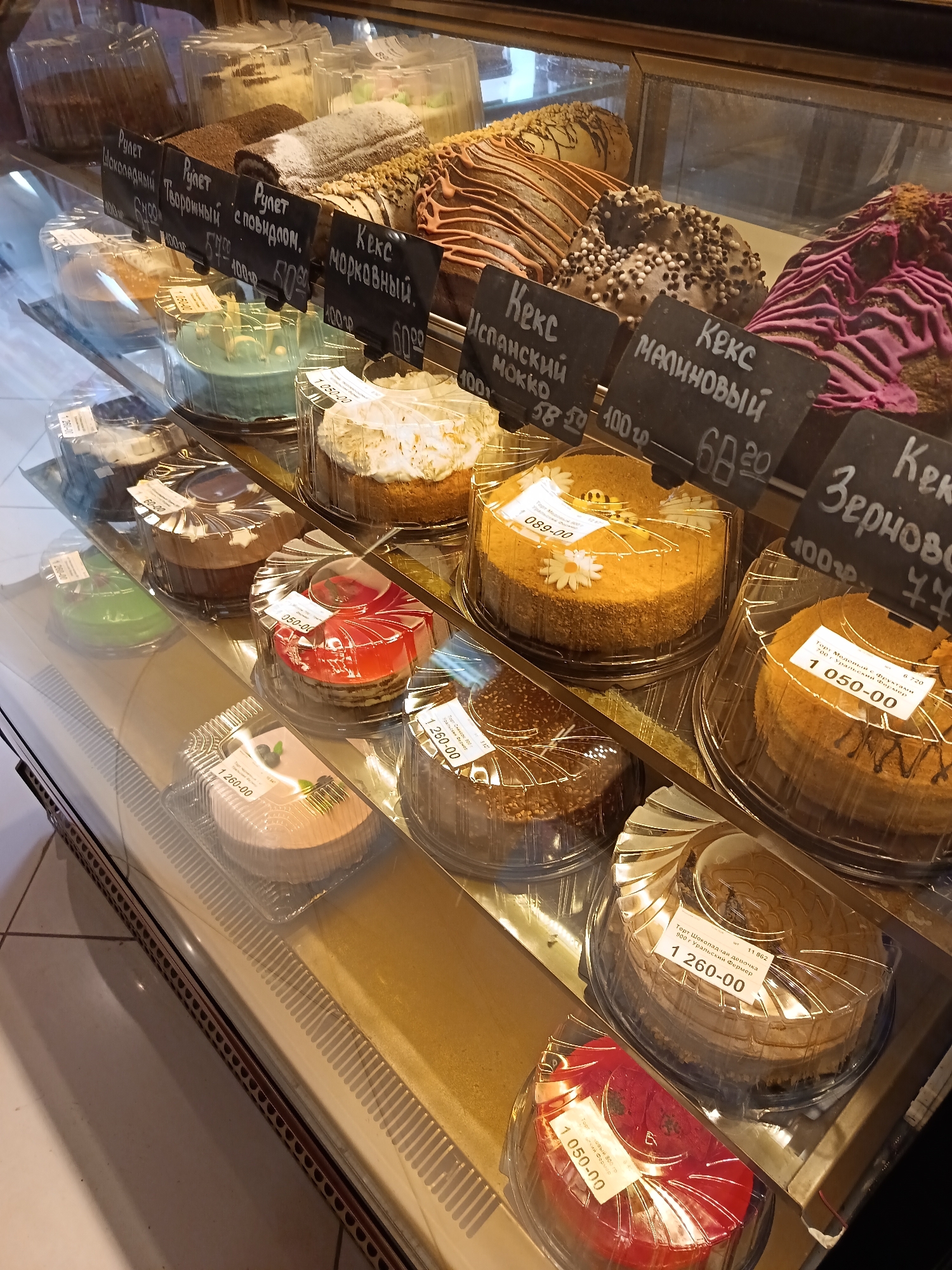 The image shows a display case filled with cakes and pastries. The items are individually packaged in clear plastic containers with price tags. The price tags indicate the names of the items in Russian, such as "Keks," "Rulet," and "Tort." The prices are listed in rubles. The items include various cakes, rolls, and other sweet treats. The display case appears to be in a bakery or cafe. The OCR data shows that some of the items are from "Ural'skiy Fermer," suggesting a local producer. The location is in the city of Osa, Russia.