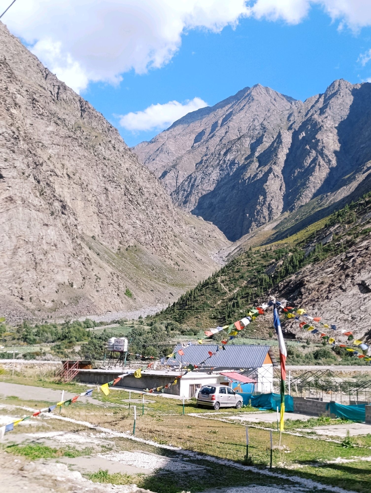 In the city of Gemur, India, this image captures a serene mountain landscape under a bright blue sky dotted with fluffy white clouds. The foreground features a small cluster of buildings with blue-tiled roofs, one of which has a water tank on its roof labeled "MUJO GUEST HOUSE." A silver car is parked in front of the building. Prayer flags, strung across the scene, flutter in the breeze, adding a vibrant splash of color. Beyond the buildings, the terrain slopes upwards, transitioning into rugged, rocky mountains. The mountains are covered in a light brown, textured surface, with deep shadows indicating their dramatic contours. Patches of green vegetation and scattered trees can be seen on the lower slopes and in the valley. The overall scene evokes a sense of peace and natural beauty, characteristic of a mountain region.