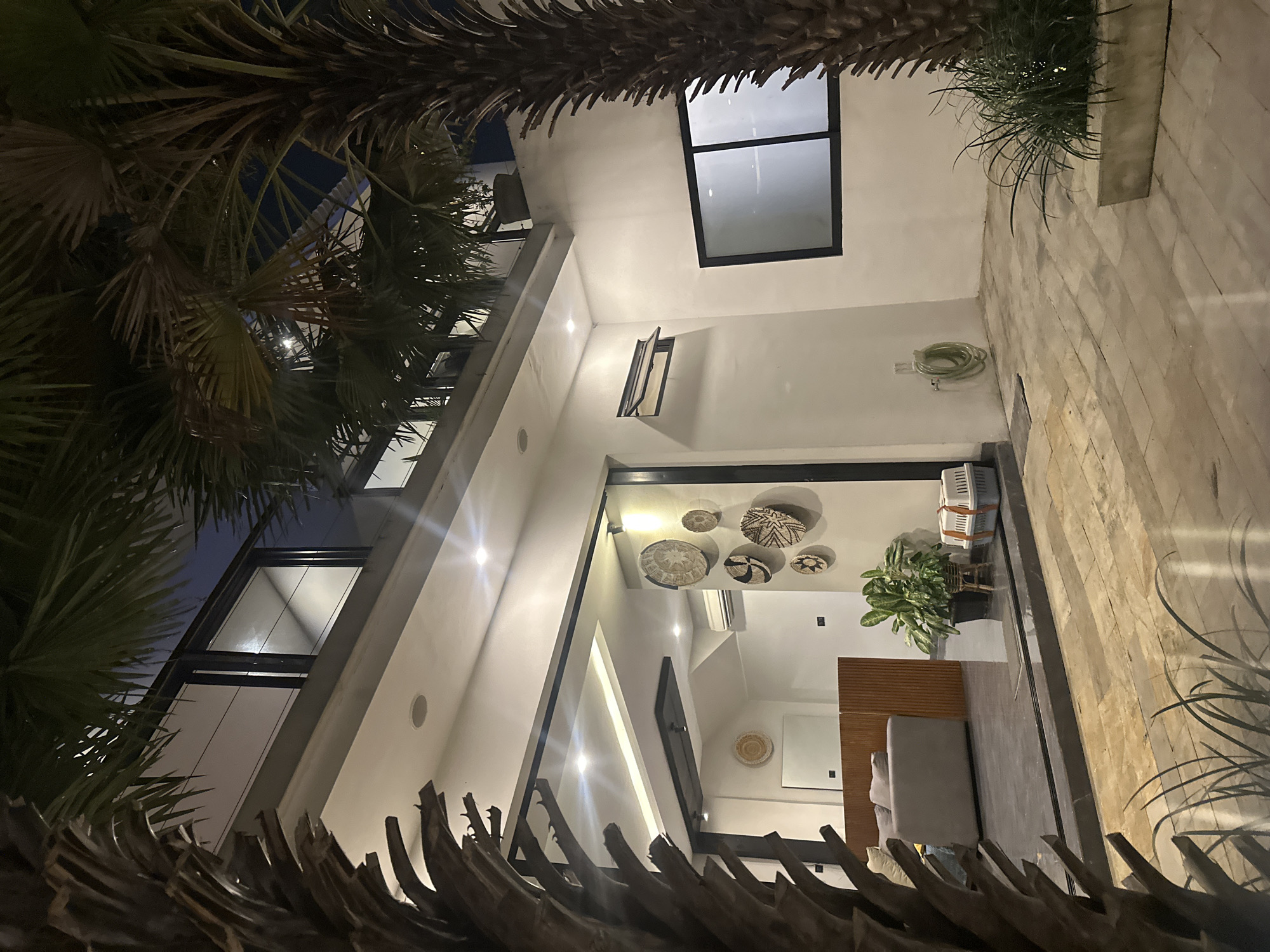 This is a night-time, low-angle, interior shot of a modern building with large windows and white walls, possibly a residential property or boutique hotel. The scene is lit by artificial lights, creating a warm glow. Lush palm fronds frame the top and left sides of the image, suggesting an outdoor, tropical environment like North Kuta, Indonesia.
Inside, the building features clean lines and minimalist decor. Several decorative woven baskets hang on the wall, adding a touch of artisanal craftsmanship. A healthy potted plant sits near a wooden partition, and a gray sofa is visible in the background, hinting at a living or lounge area. On a shelf above the plant, two white pet carriers are stacked. The floor is paved with large, rectangular light-colored stones. A coiled green hose is attached to the wall near a small, open window. The overall impression is one of sophisticated, contemporary design in a warm, possibly humid, climate. The image does not show any people or visible text.