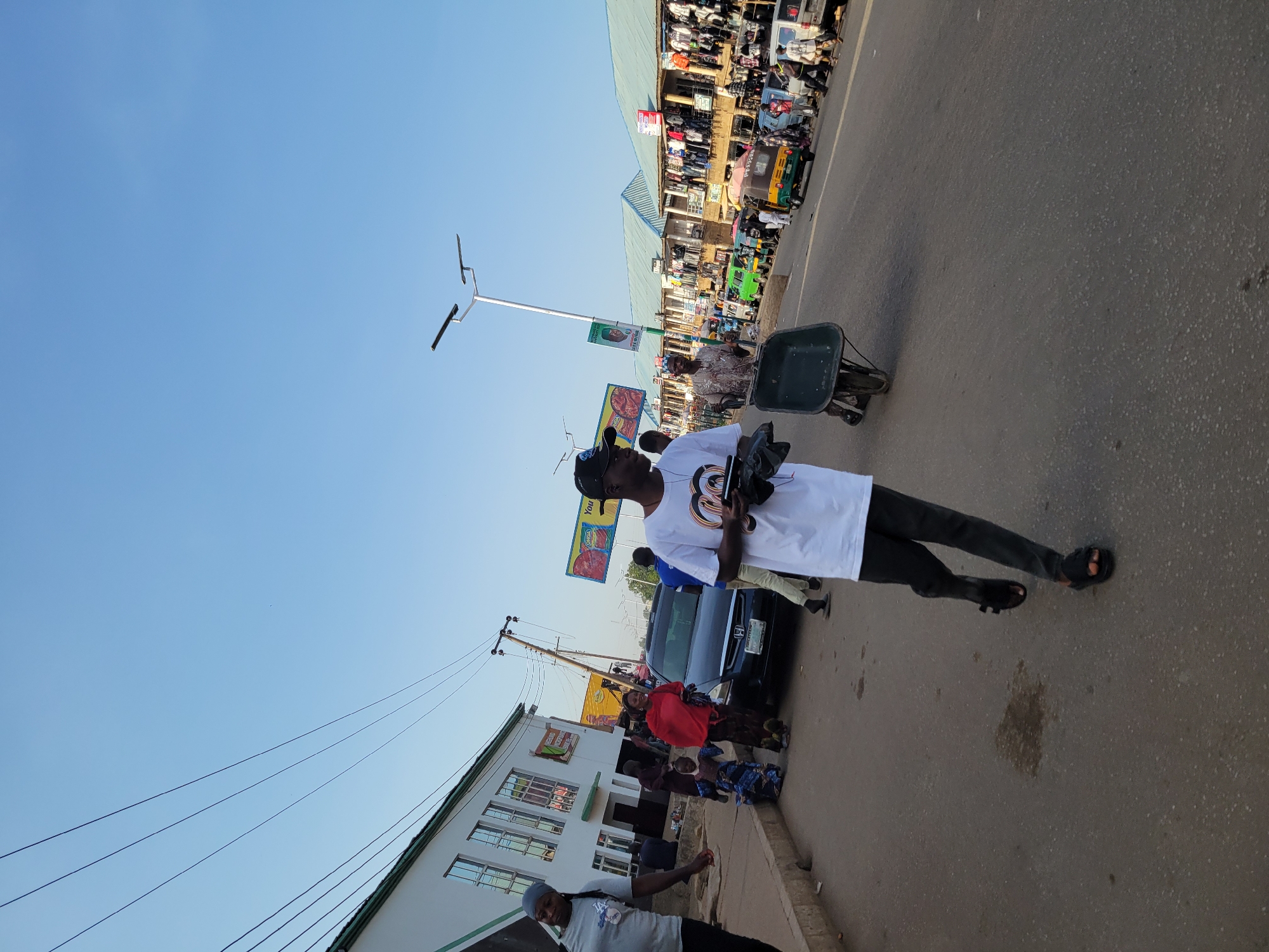 A street in Bukuru, Nigeria, features a man wearing a white t-shirt and black pants, holding an object and walking on the right side of the road. There are several other people on the road, some standing by a parked black car. A wheelbarrow stands nearby. On one side of the road is a building with multiple windows, and on the other side, shops and people are visible under a covered market structure. Overhead power lines are visible. The sky is clear and blue.