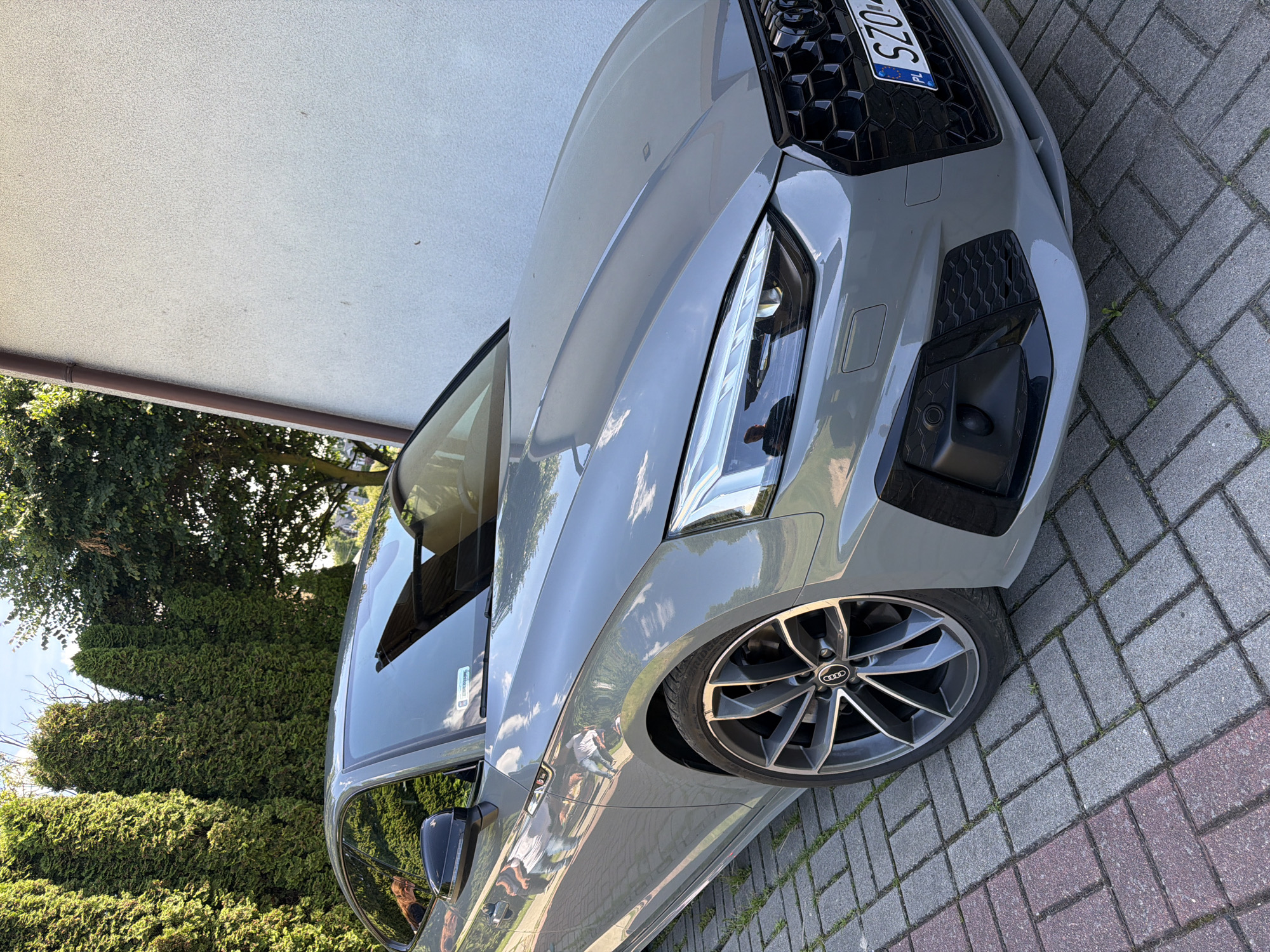 The image depicts a grey Audi A5 coupe parked on a paver driveway in front of a white stucco building. The car's front right wheel is prominently featured, and the vehicle's sleek design is evident. In the background, tall green coniferous trees line the property, suggesting a suburban or rural setting. A glimpse of the sky indicates it is daytime. The car has a Polish license plate that reads "SZO...". The overall scene is static, with no people or activity visible.