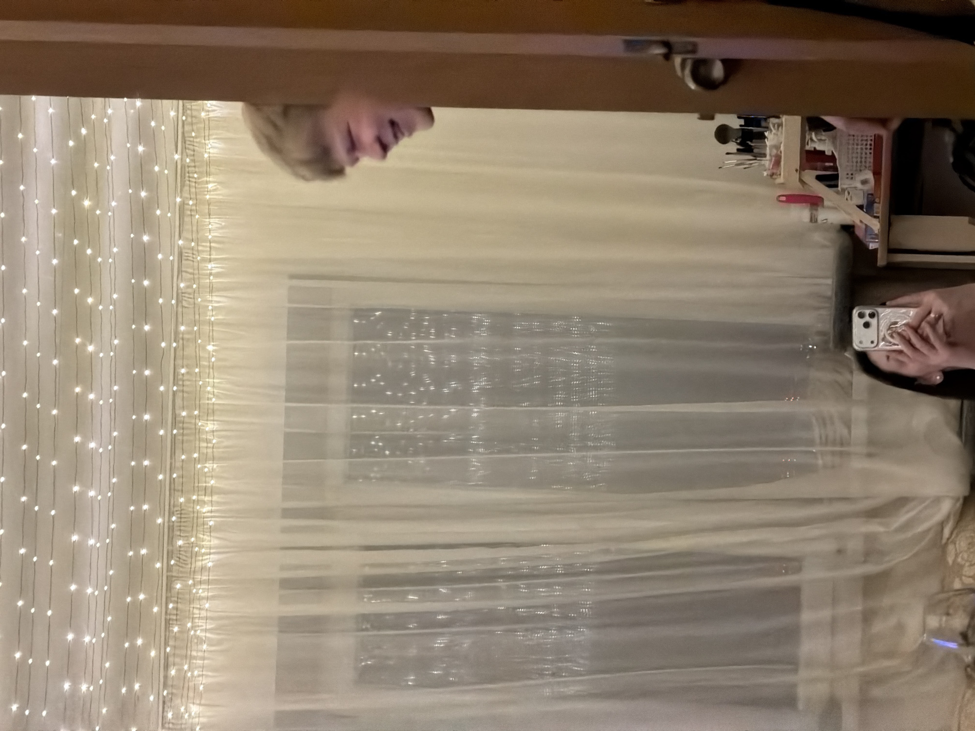 A reflection in a mirror reveals a room interior. The mirror is framed by a wooden edge, with a section of a person's face visible at the top. Below, a second person holds a smartphone, capturing the reflection. In the background, a window is covered by a sheer white curtain adorned with strands of small fairy lights. A shelf with various items including makeup brushes and other cosmetic tools is visible. The location is likely indoors, but there are no external details to confirm if it is in Izhevsk, Russia.