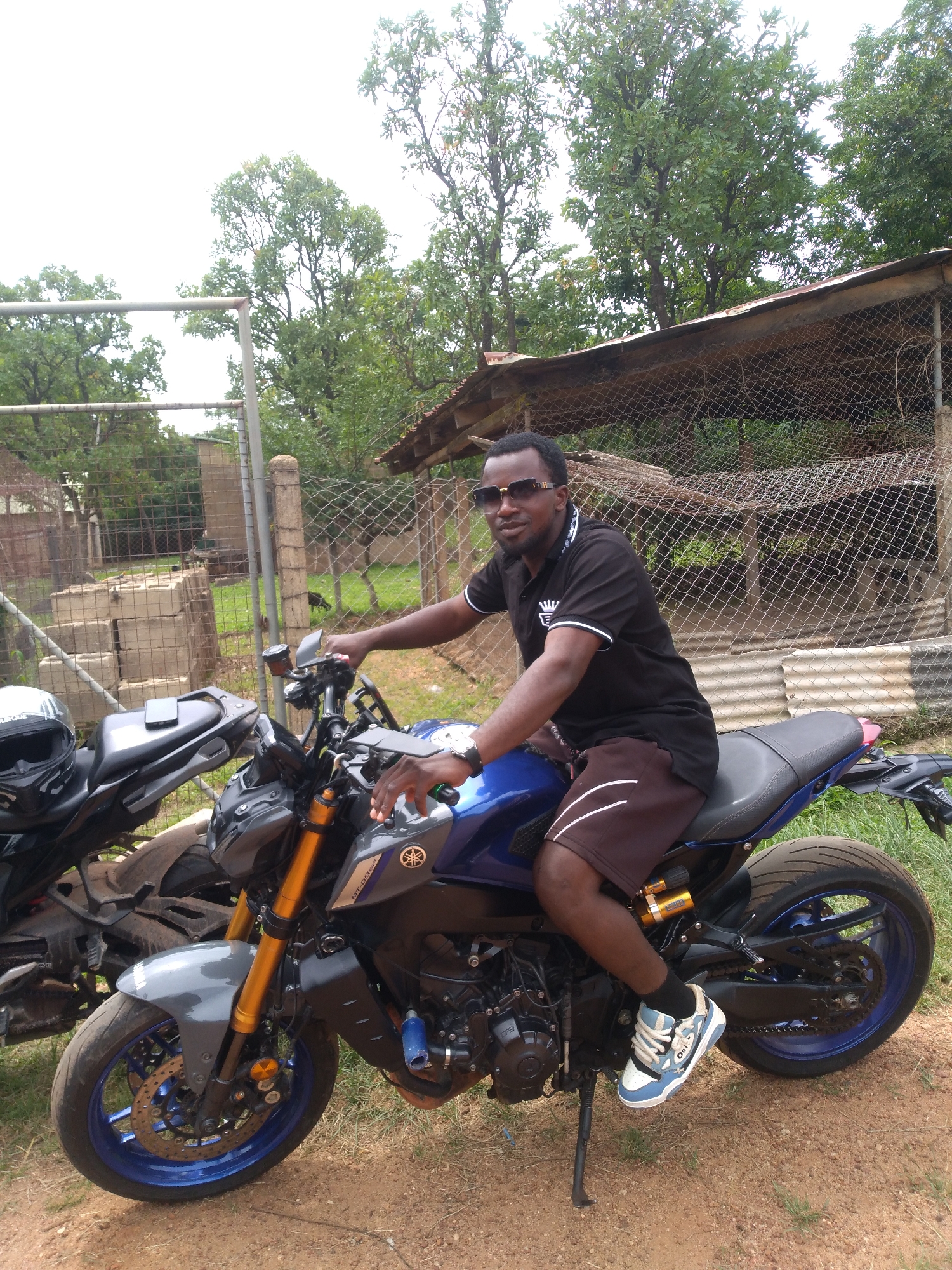 A man, appearing to be in his late 20s or early 30s, sits astride a blue and gray Yamaha motorcycle. He is wearing a black polo shirt with white trim and brown shorts. His feet are clad in light blue and white sneakers. The man has dark skin, short black hair, and is wearing gold-rimmed sunglasses. He is positioned in the foreground of the image. Behind him, another dark-colored motorcycle is partially visible. The background consists of a chain-link fence, some trees, and what appears to be a rustic structure with a corrugated metal roof. The ground is a mix of dirt and sparse grass. The setting suggests an outdoor area, possibly a residential compound or a rural property. The location is identified as Jos, Nigeria.